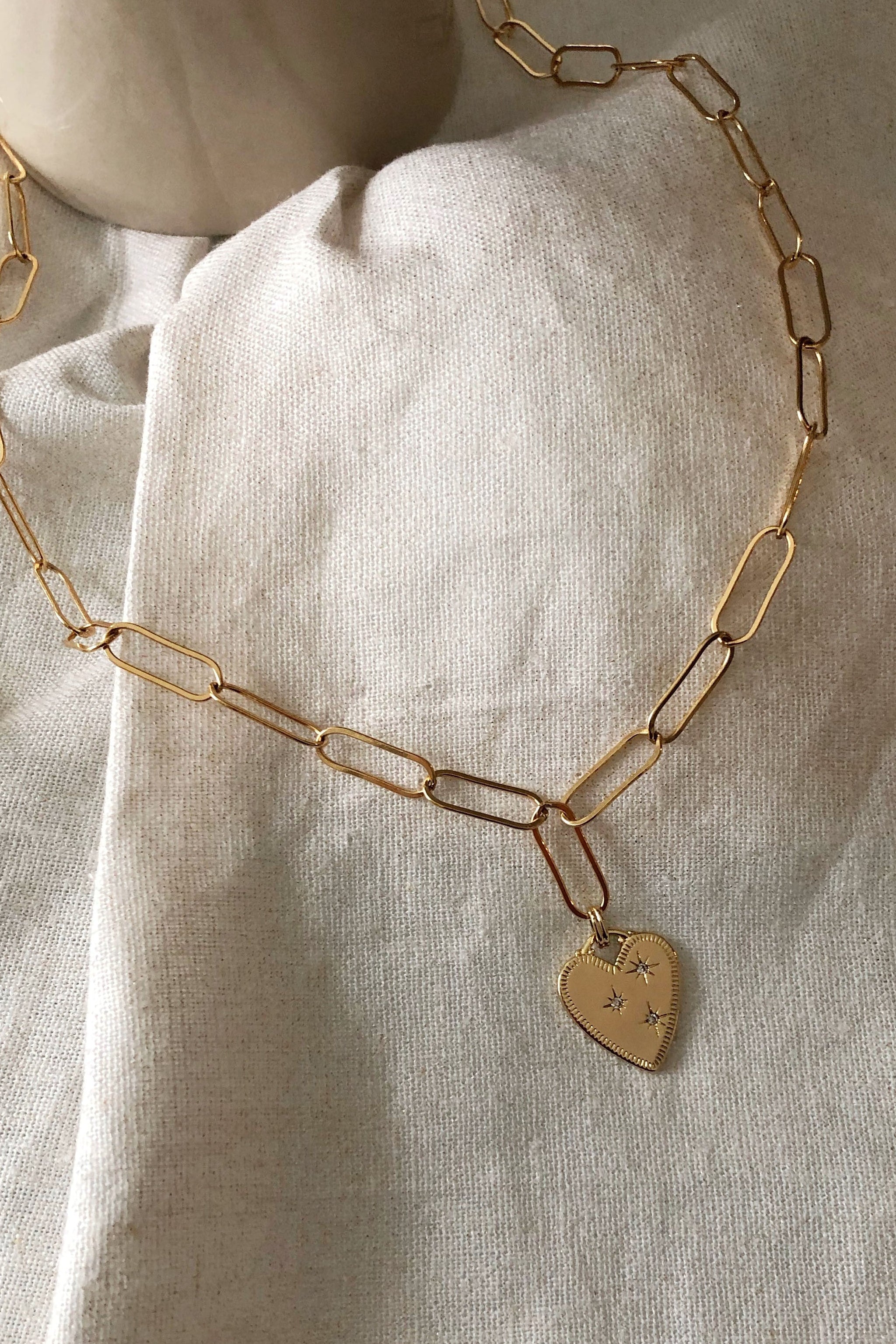 Gold chain necklace with a heart-shaped tag on a light fabric background with white flowers.