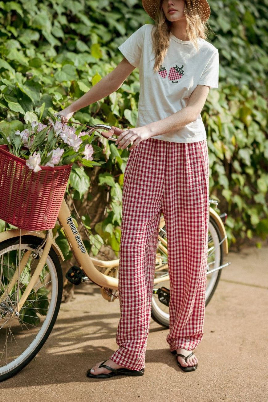 Woman in a white t-shirt and red checkered pants standing next to a bicycle with a basket, against a green leafy background.