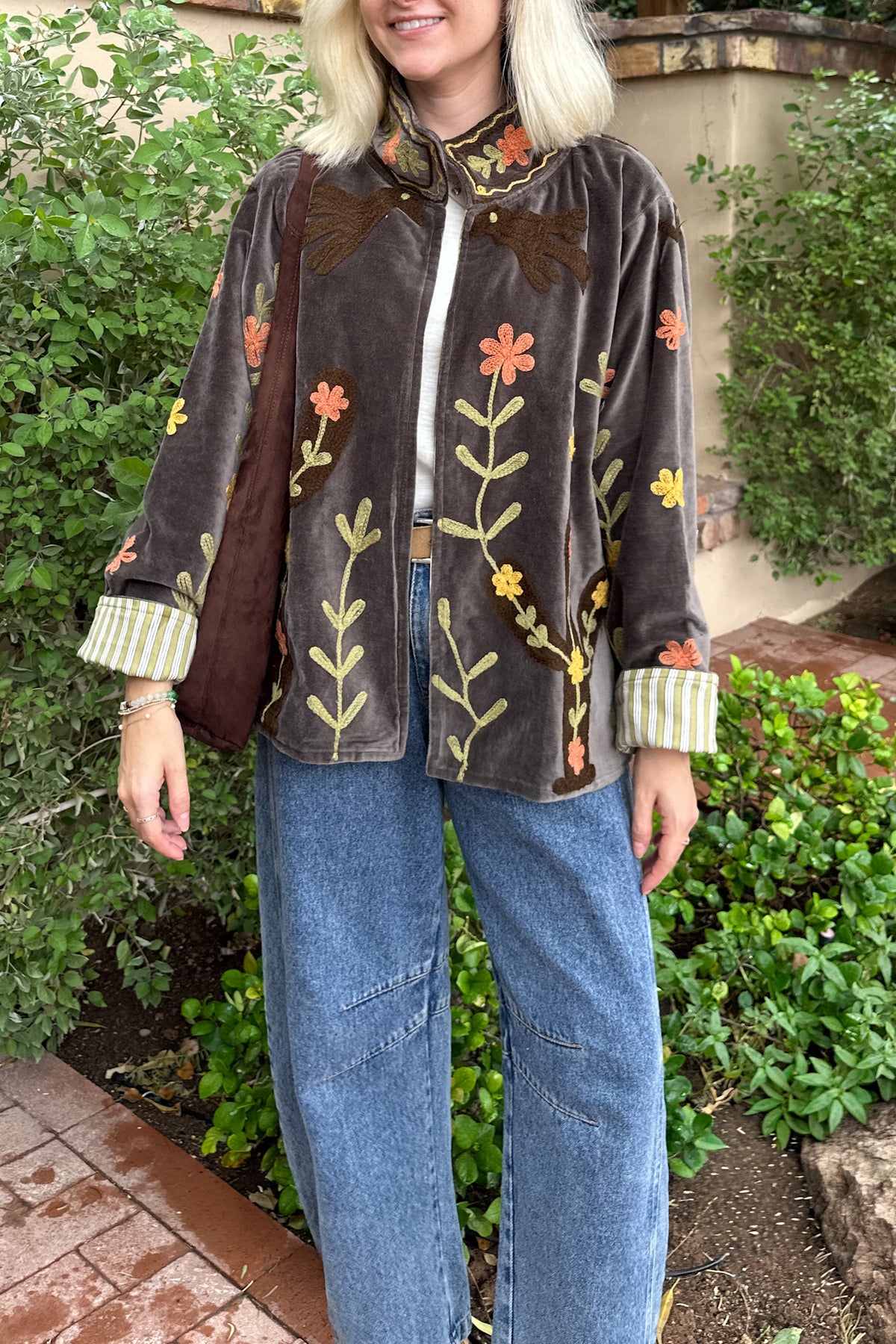 A woman wearing a brown velvet jacket with colorful floral embroidery, blue wide-leg jeans, and a striped shirt, standing outdoors in front of green bushes.