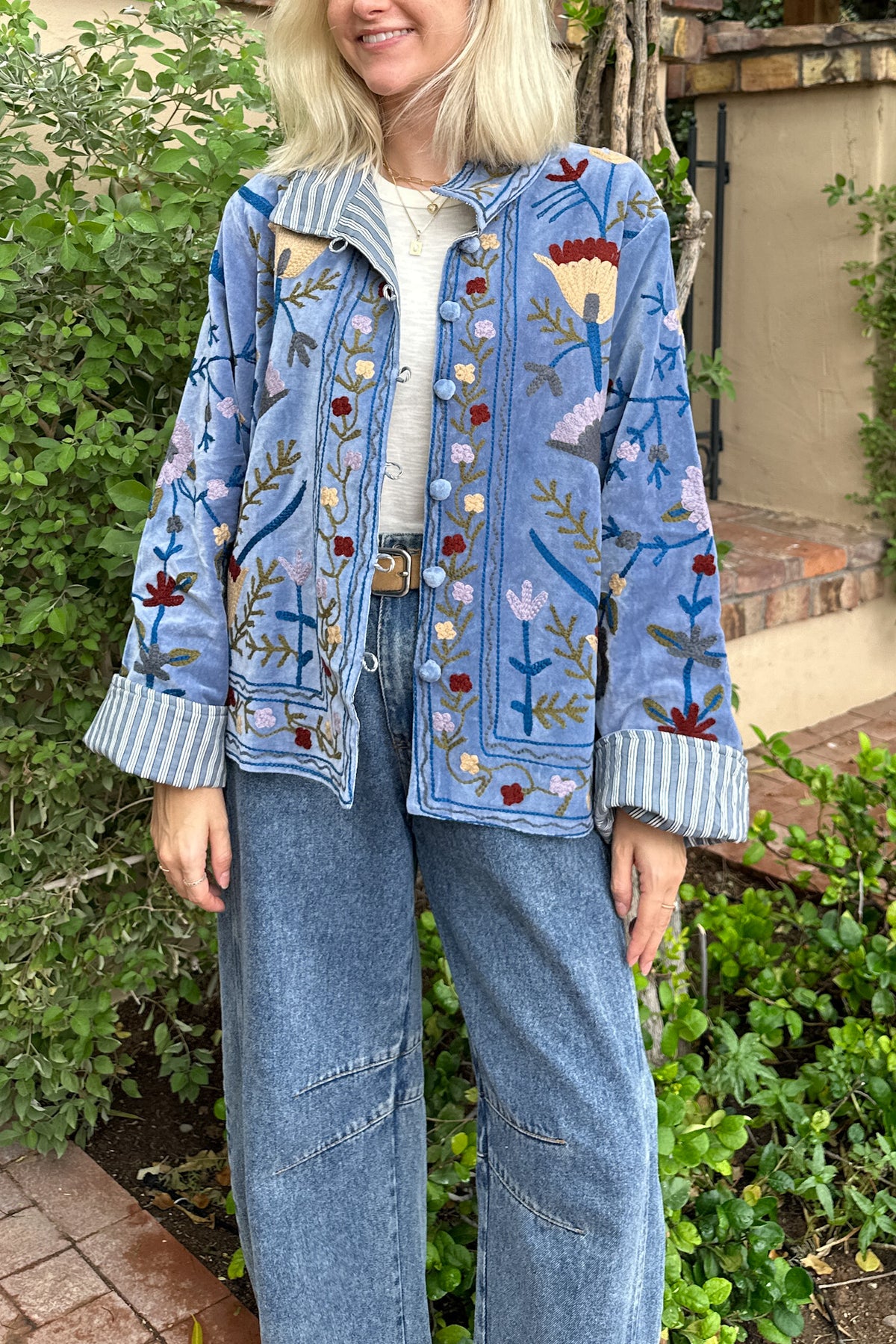 A woman wearing a light blue denim jacket with colorful floral embroidery, striped button-up cuffs, and a white top, paired with wide-leg blue jeans, standing outdoors in front of green foliage.