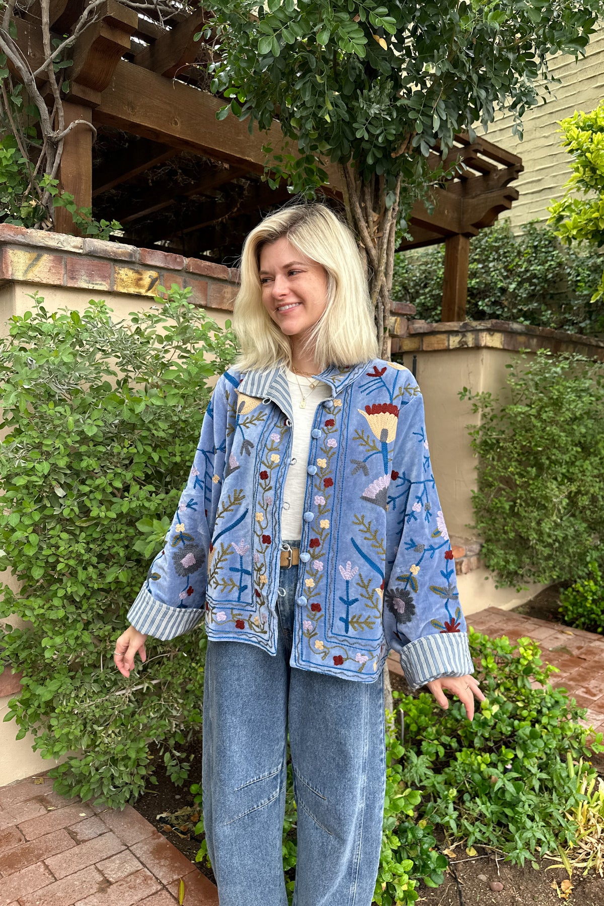 A woman wearing a light blue denim jacket with colorful floral embroidery and striped button details, paired with light blue jeans, standing in a garden setting.