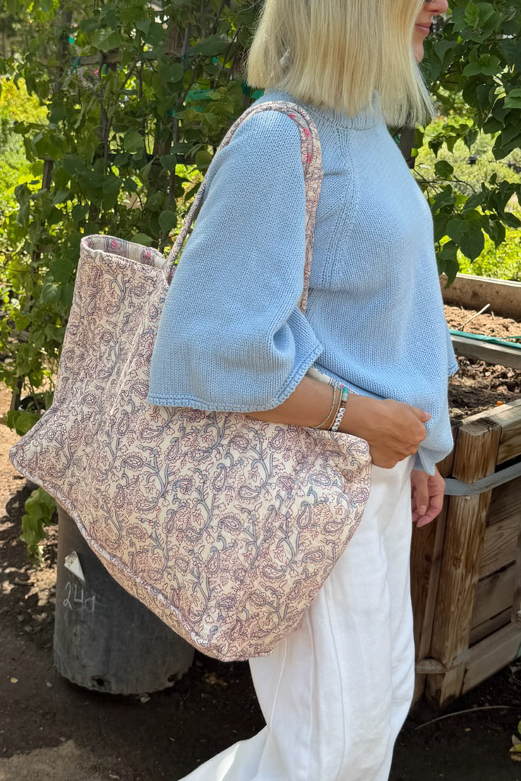 A woman outdoors wearing a light blue knit sweater and white pants, carrying a large soft tote bag with a beige, pink, and blue paisley pattern.