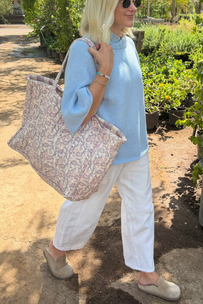 A woman walking outdoors in a garden center carrying a large patterned tote bag over her shoulder. She is wearing a light blue knit sweater, white wide-leg pants, and beige slip-on shoes.