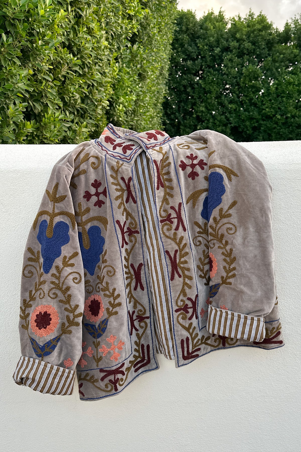 A vintage-style embroidered jacket or cardigan in taupe velvet, featuring colorful floral and bird motifs in blue, pink, and brown, draped against a white wall with green hedges in the background.