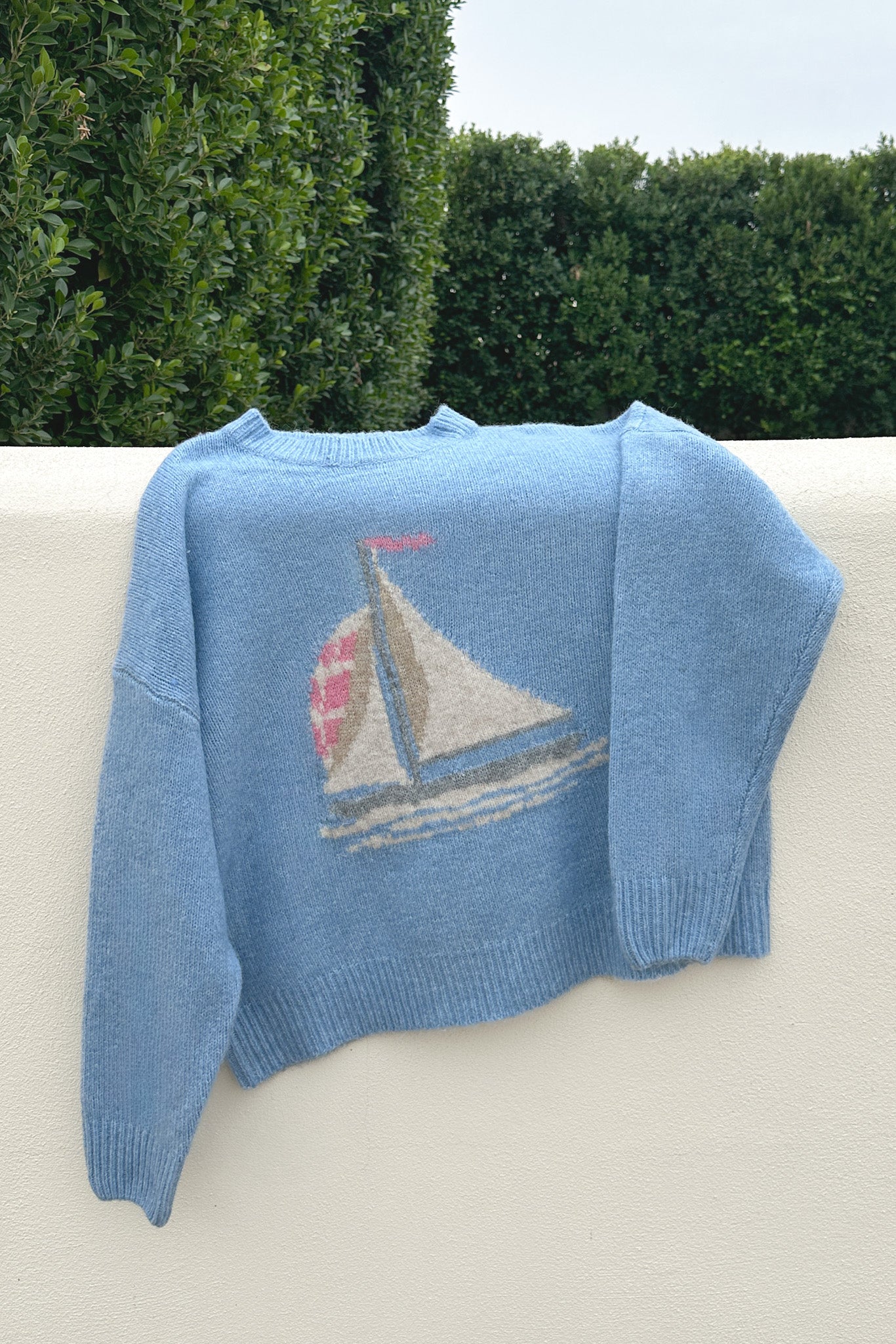 Blue knitted sweater with a sailboat design on a white surface with greenery in the background