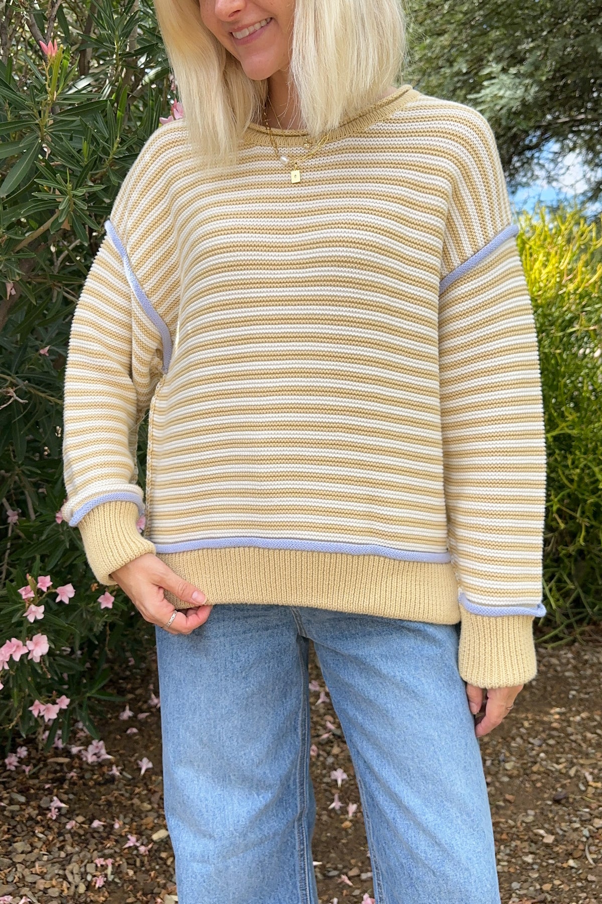 A woman wearing a cream and tan striped knit sweater with lavender trim, paired with light blue jeans, standing outdoors in front of green bushes.