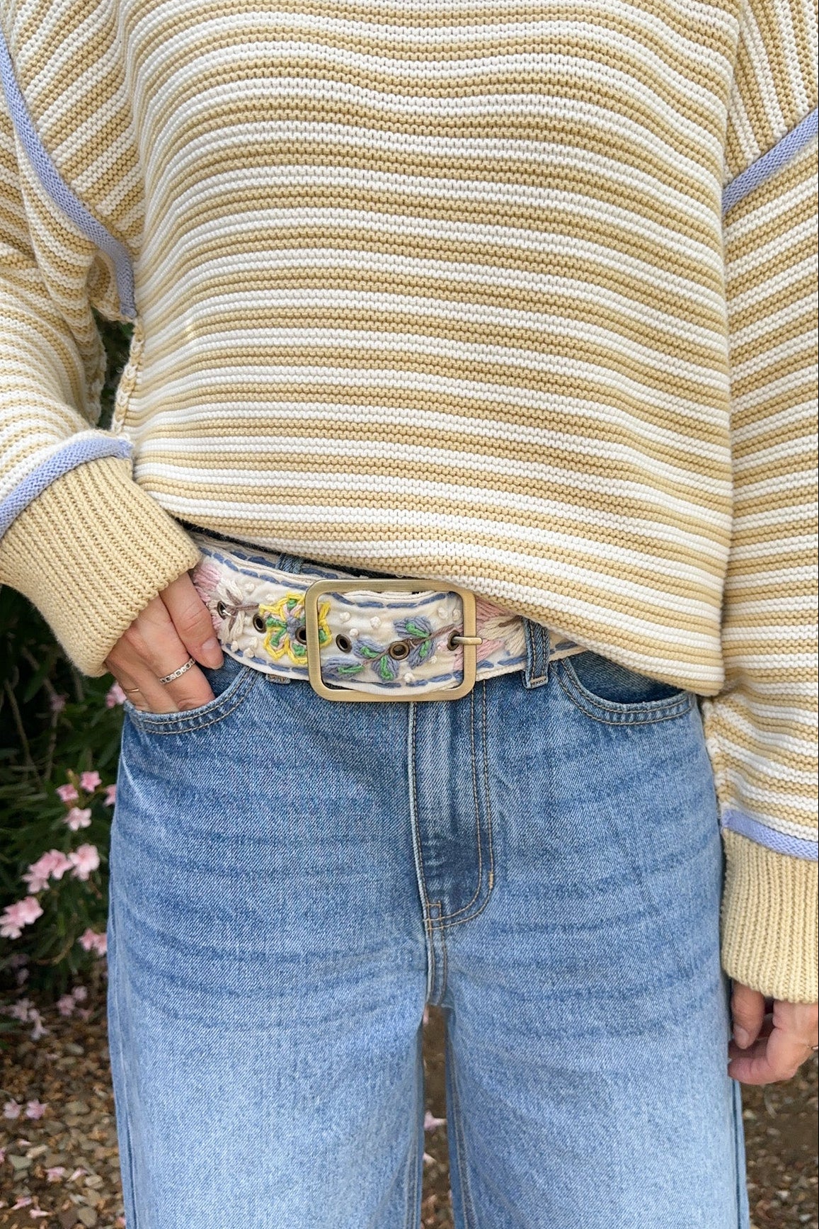 A person wearing a cream and tan striped knit sweater with light blue ribbed trim, tucked into light-wash blue jeans with a decorative belt, photographed outdoors in a garden setting.