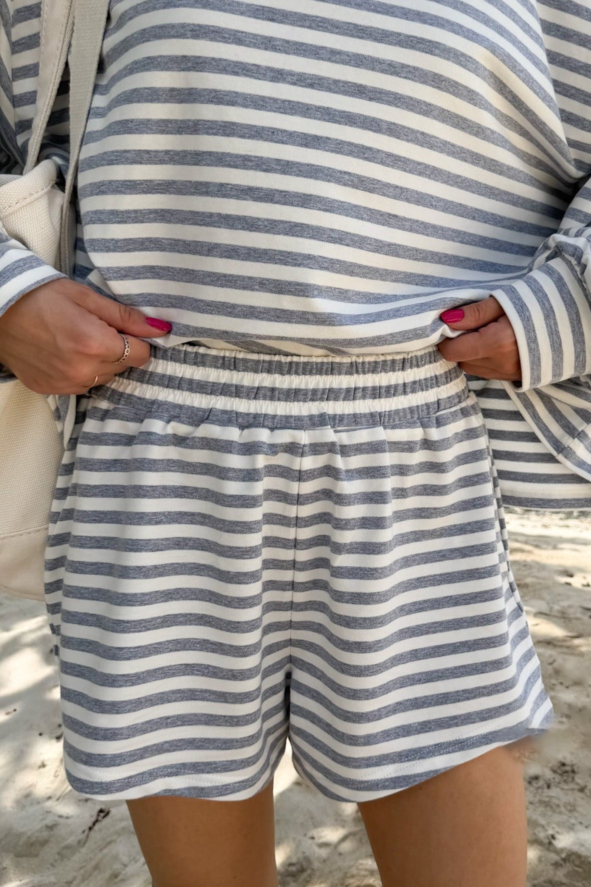 A person standing on a sandy beach wearing a matching long-sleeve top and shorts set with horizontal blue-gray and off-white stripes. The person is holding the waistband of the shorts, with a cream shoulder bag visible on the left side.