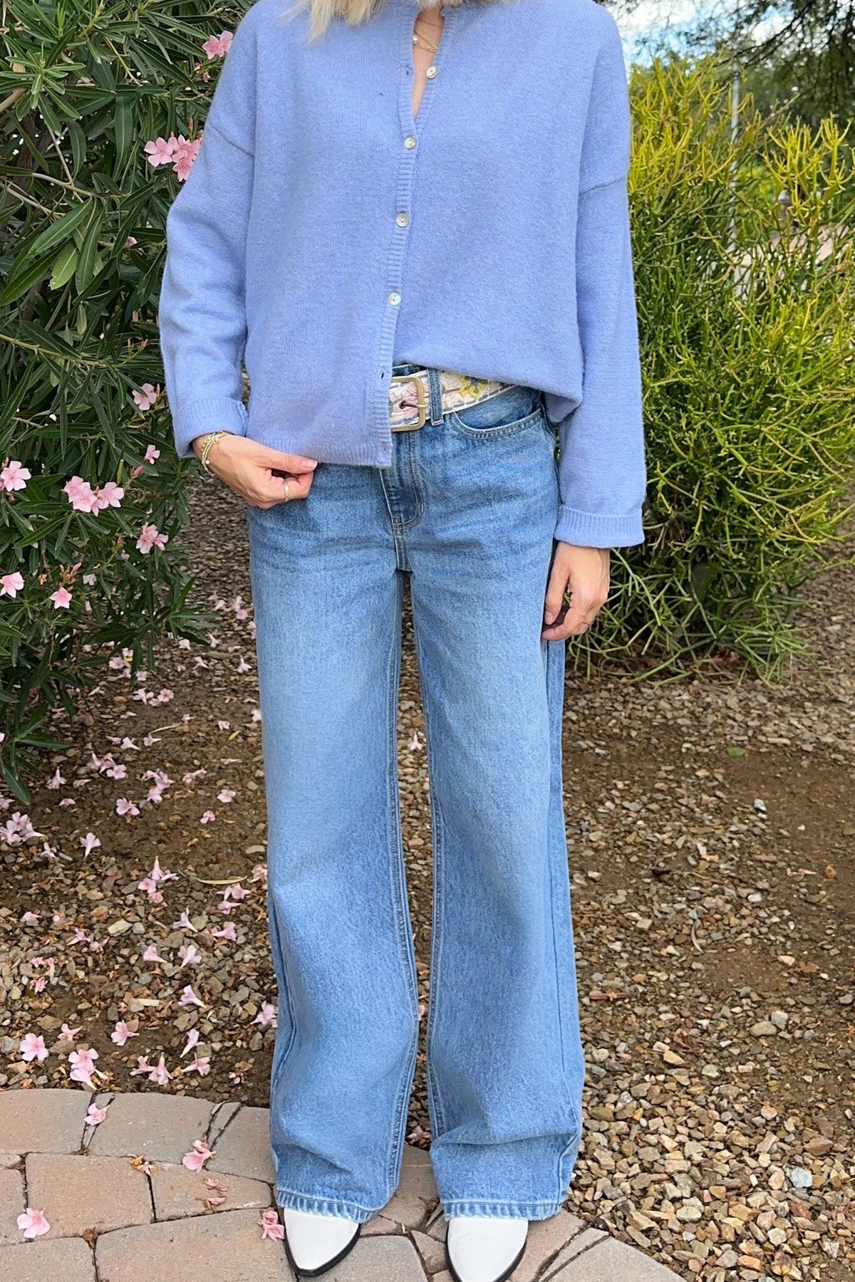 A woman wearing a light periwinkle blue cropped button-up knit cardigan and wide-leg light blue jeans, standing outdoors in a garden setting.