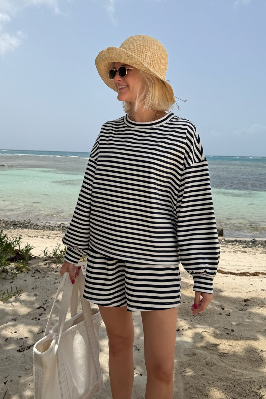 A woman stands on a sandy beach wearing a navy and white horizontally striped long-sleeve top and matching shorts, a straw hat, and holding a white tote bag.