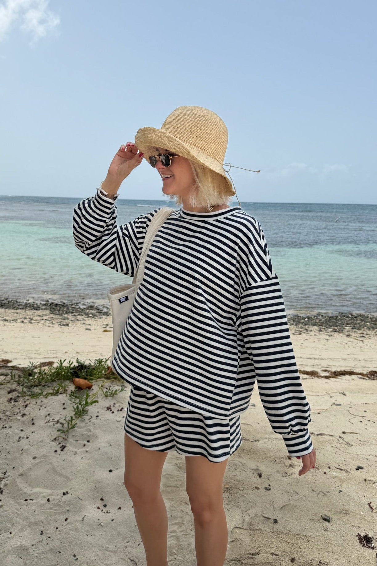 A woman stands on a sandy beach wearing a navy and white horizontally striped long-sleeve top and matching shorts, a beige straw sun hat, and a white shoulder bag, with the ocean and blue sky in the background.