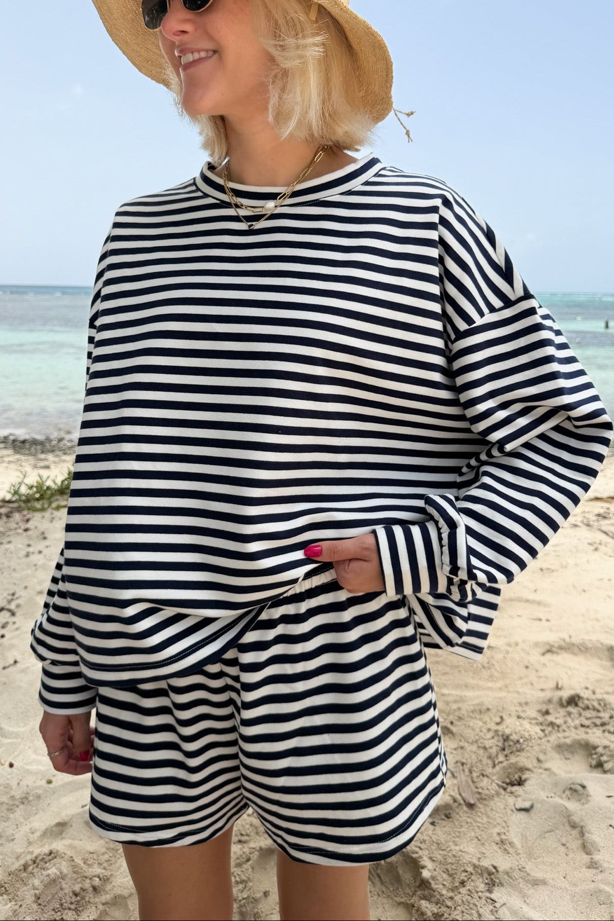 A woman standing on a sandy beach wearing a matching navy and white horizontally striped long-sleeve top and shorts set, a straw hat, and sunglasses.