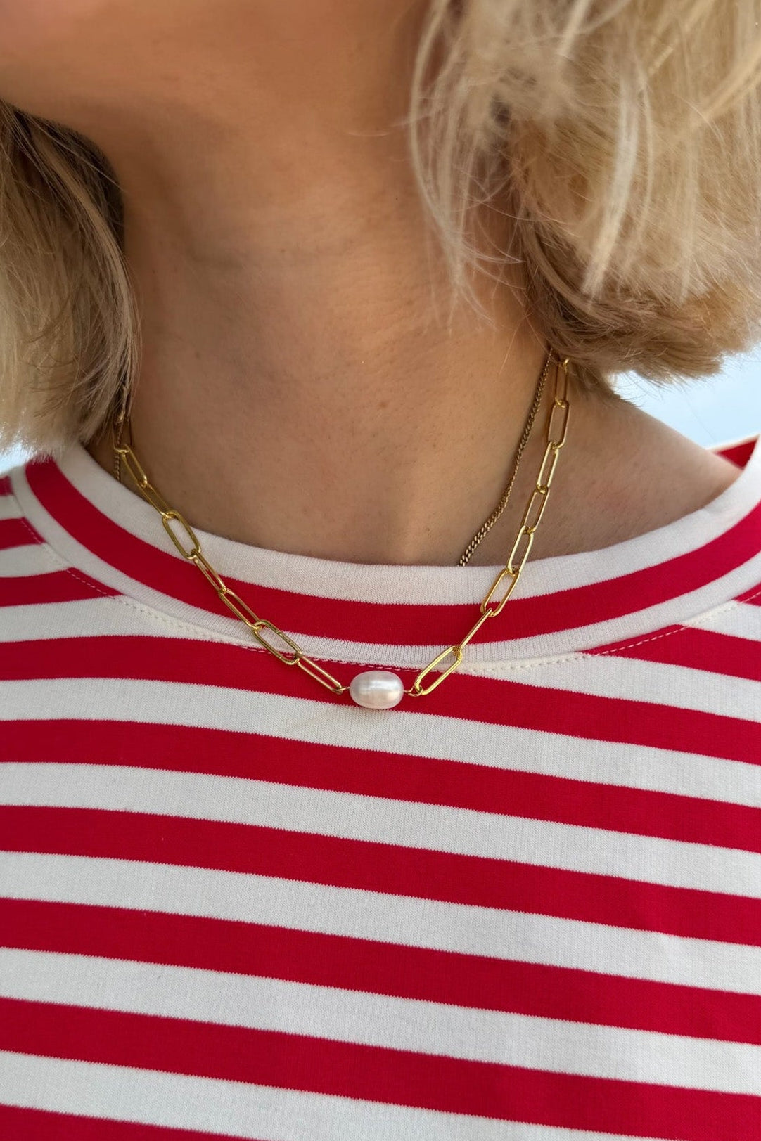 A close-up of a person wearing a delicate gold chain necklace with a single white pearl pendant, paired with a red and white striped shirt.