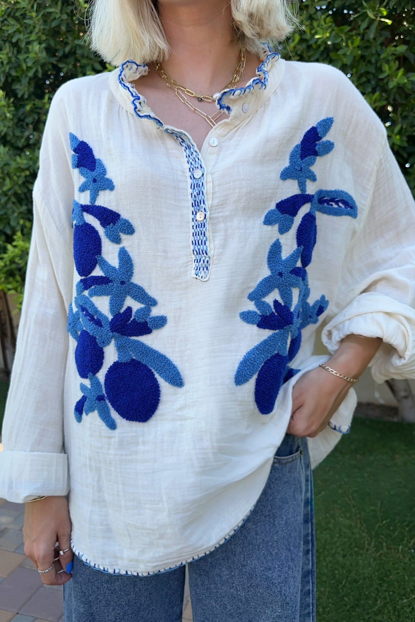 A woman wearing a loose white linen top with blue floral embroidery and a ruffled collar, paired with blue jeans, photographed outdoors in front of green foliage.
