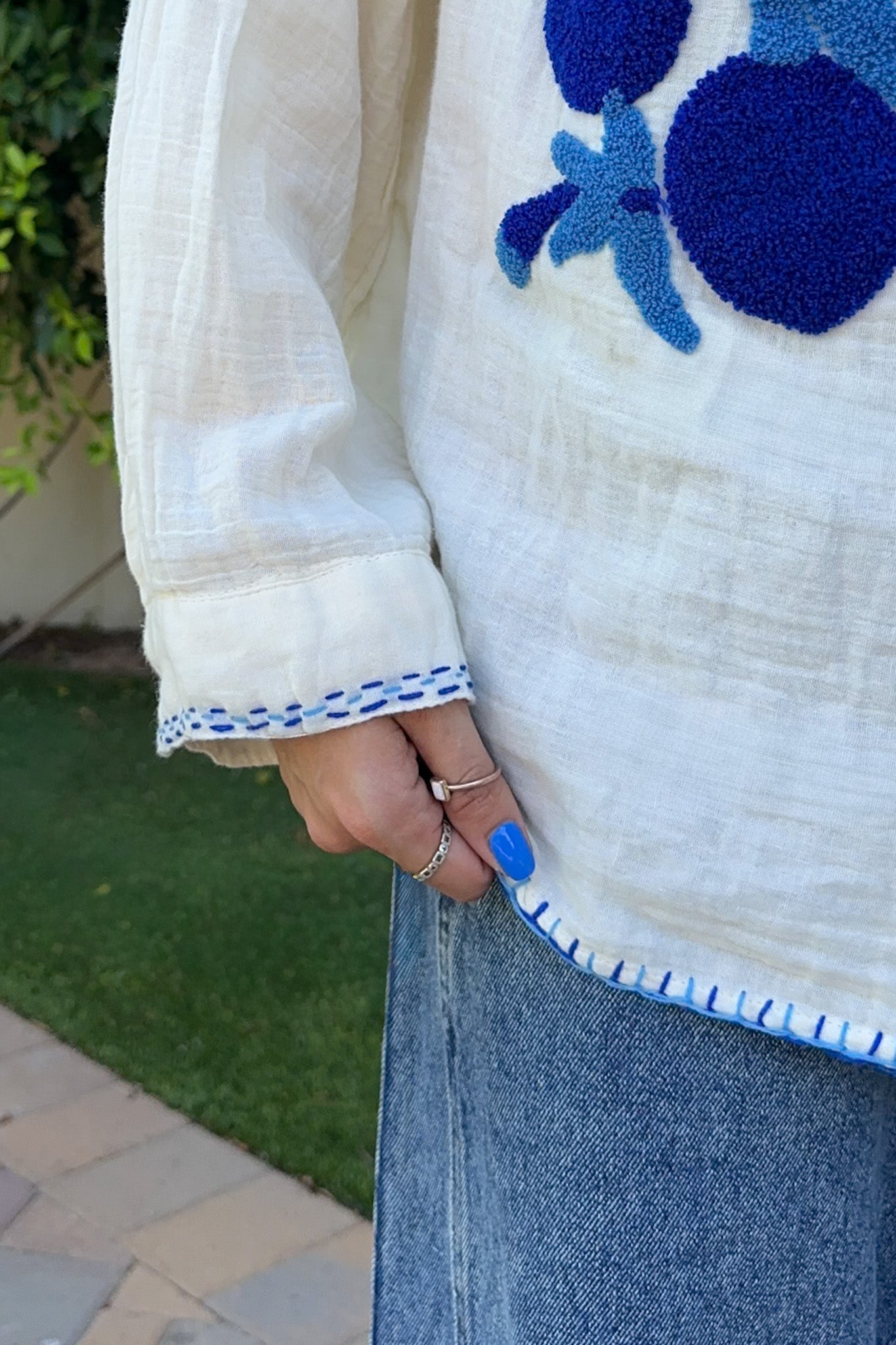 A close-up of a person wearing a white textured blouse with blue embroidered floral details and blue-and-white striped trim on the cuffs and hem, paired with a blue denim skirt. The photo is taken outdoors with grass and paving stones visible in the background.