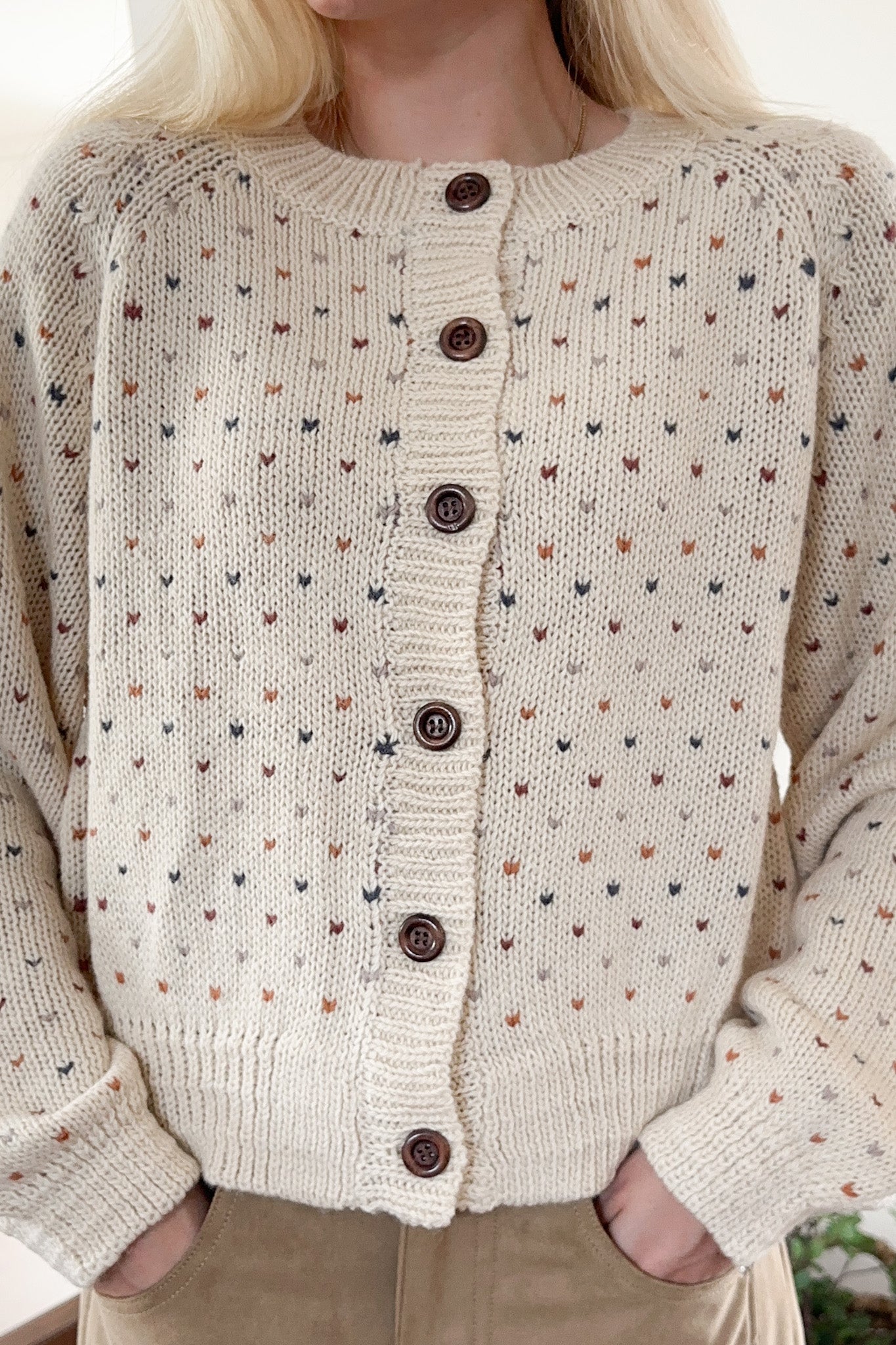 Close-up of a person wearing a beige knitted cardigan with small patterns and brown buttons.