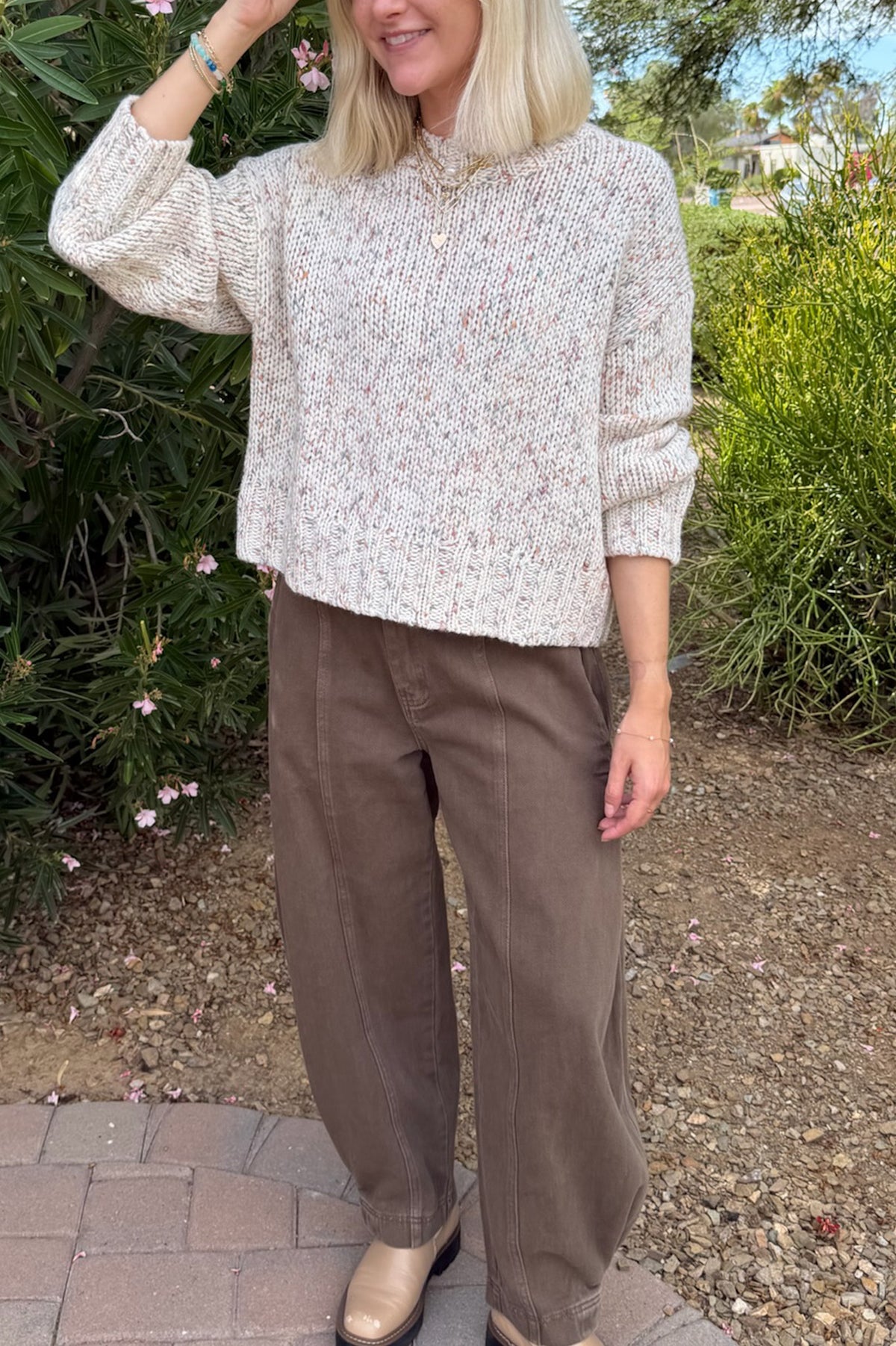 A woman wearing a cream-colored, textured knit mock-neck sweater with subtle multicolor flecks, paired with brown wide-leg pants and beige boots, standing outdoors in front of green garden foliage.