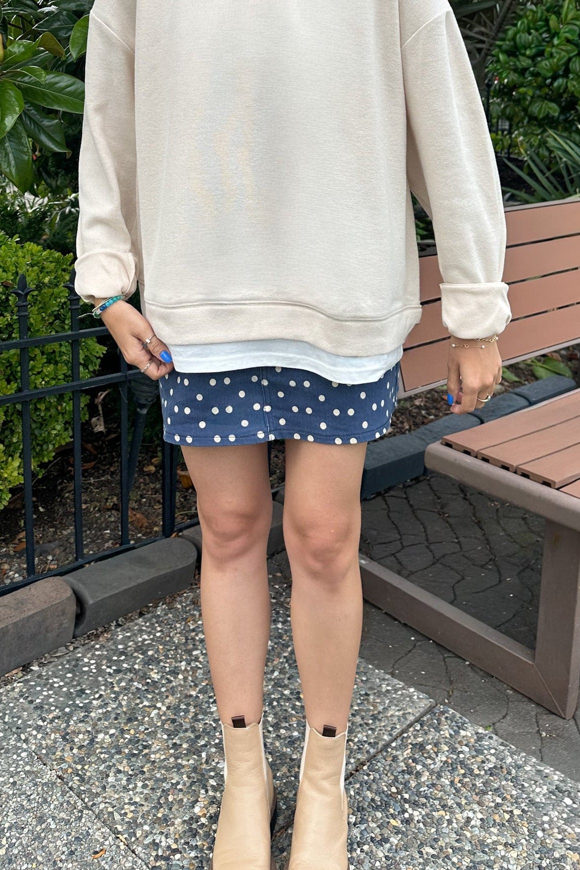 A person standing outdoors wearing an oversized cream sweatshirt, a navy blue mini skirt with white polka dots, and beige ankle boots, with green foliage and a wooden bench in the background.