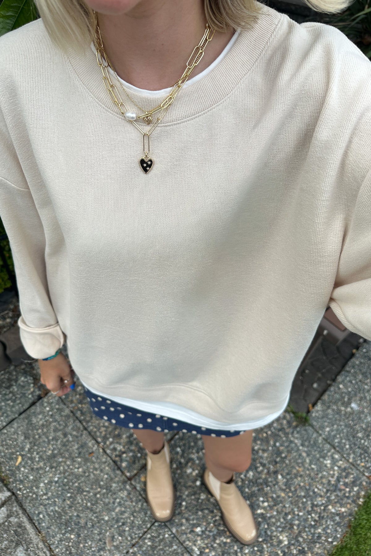 A person wearing an oversized cream-colored ribbed sweatshirt, layered with gold chain necklaces, a navy blue polka-dot skirt, and beige ankle boots, standing outdoors on a paved surface.