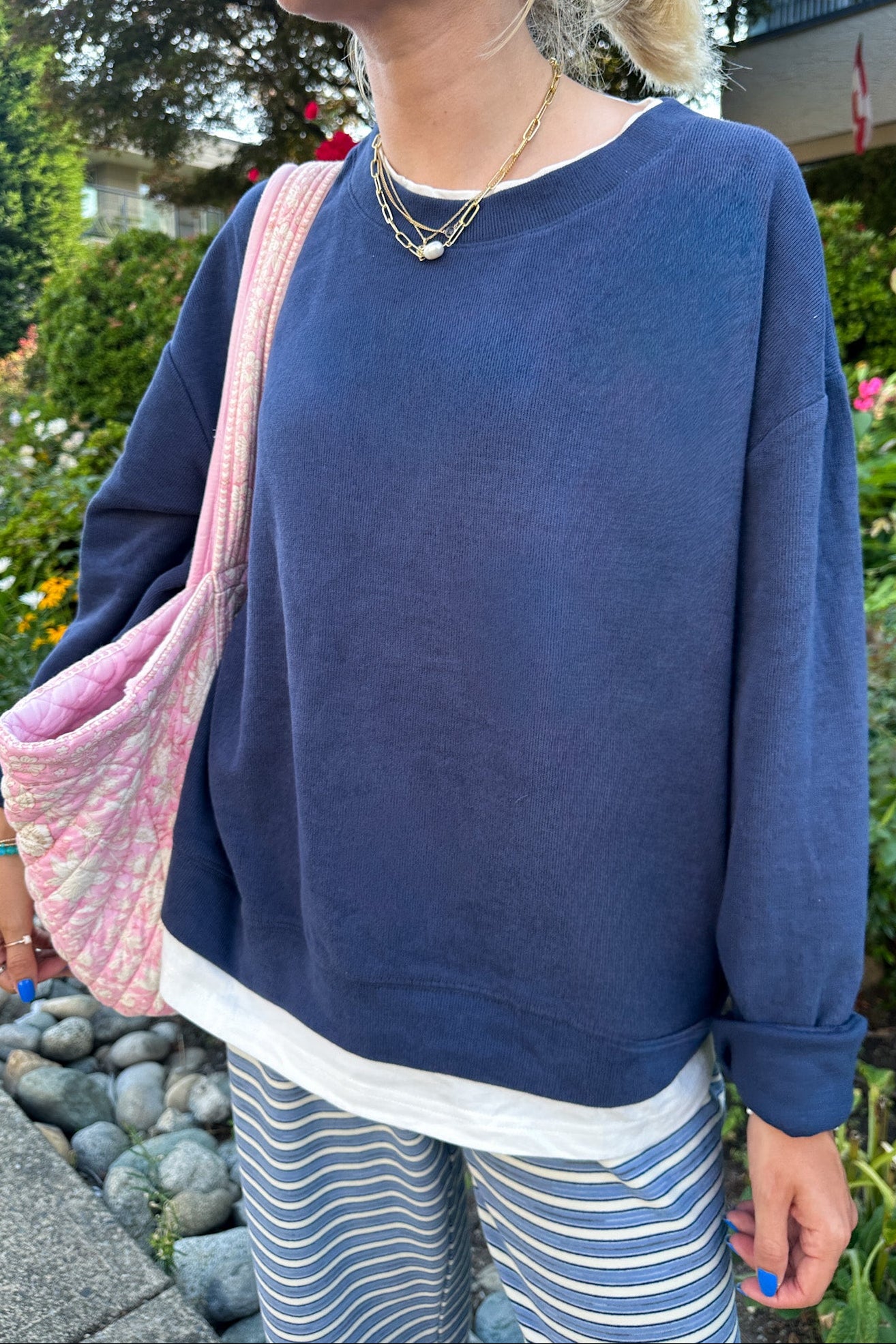 A person outdoors wearing an oversized navy blue sweatshirt with a white hem, striped pants, and a pink patterned shoulder bag.