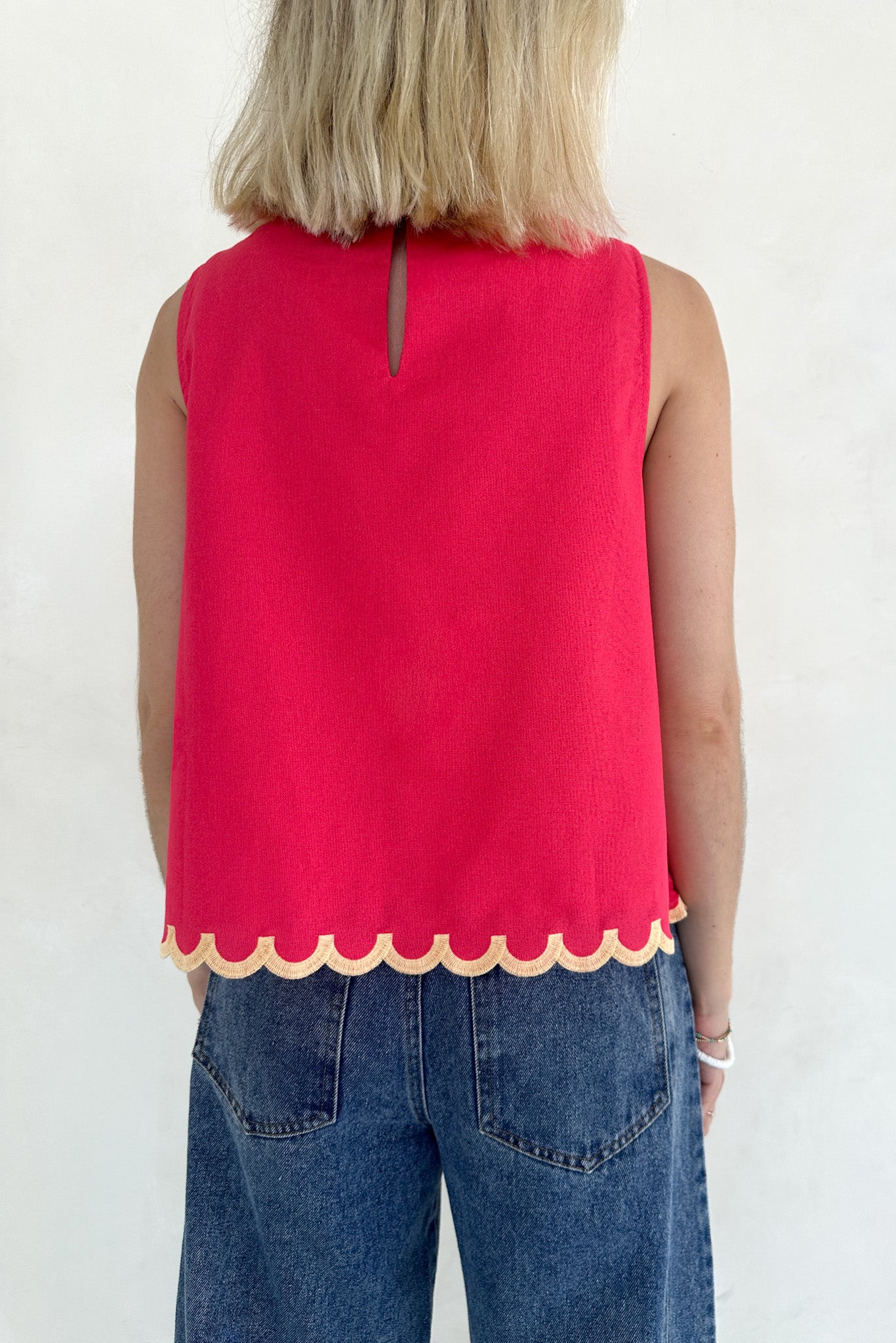 Person wearing a red sleeveless top with scalloped hem and blue jeans on a white background