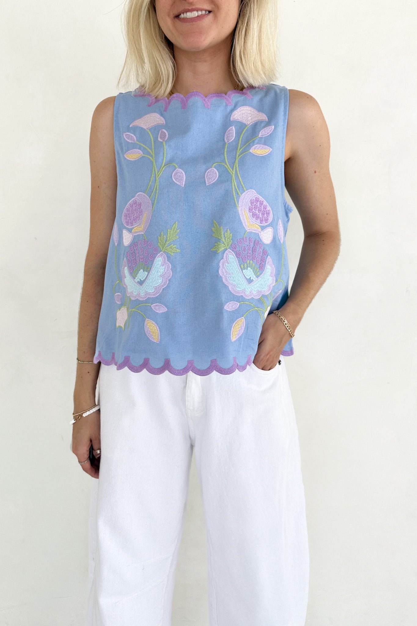 Woman wearing a light blue sleeveless top with floral patterns and white pants on a plain background
