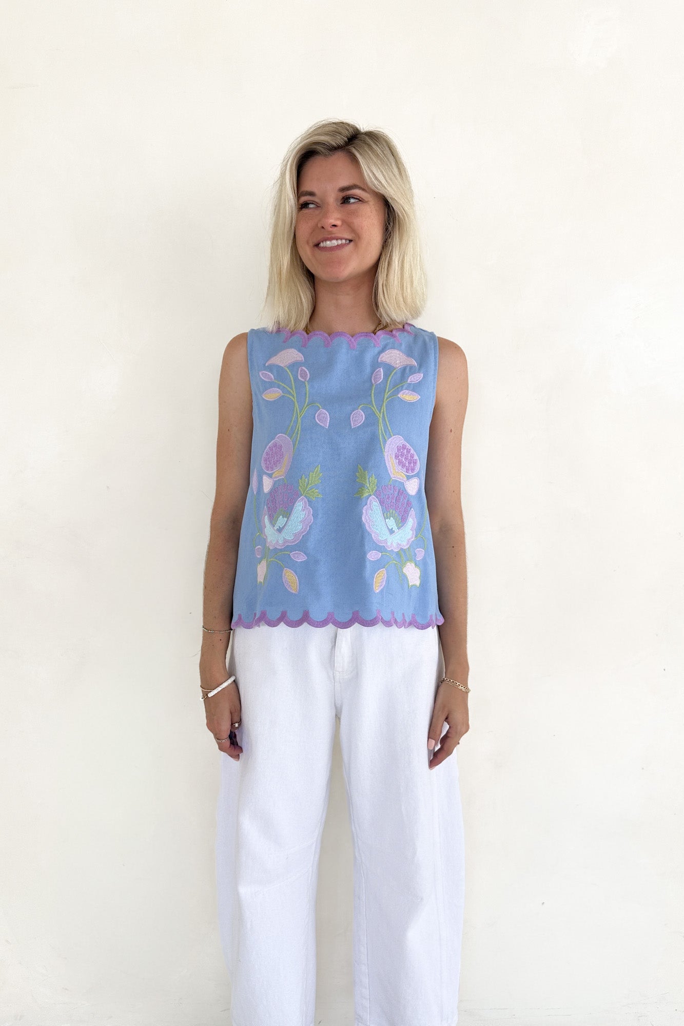 Woman wearing a blue sleeveless top with floral embroidery and white pants on a white background