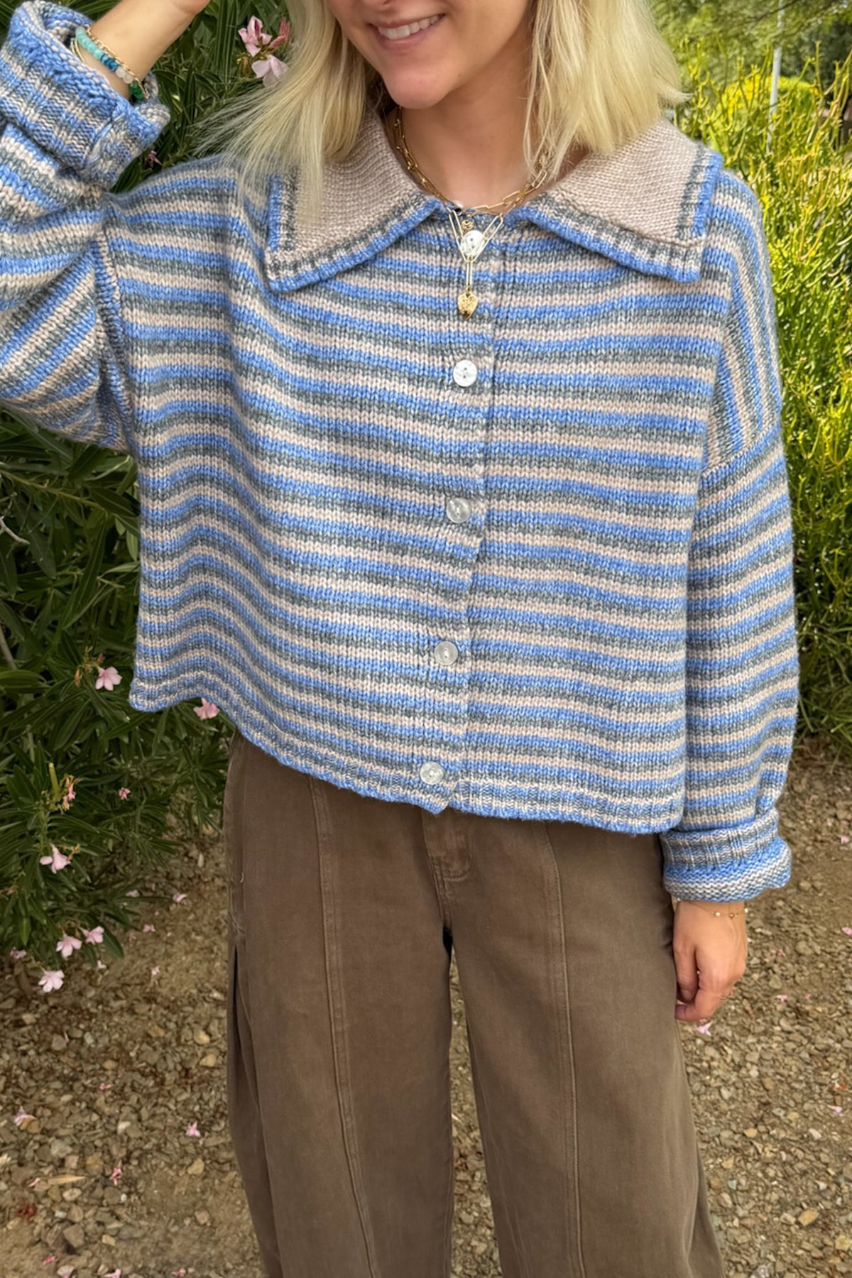 A woman wearing a blue and gray striped knit cardigan with a beige collar, paired with brown wide-leg pants, standing outdoors in front of green foliage.