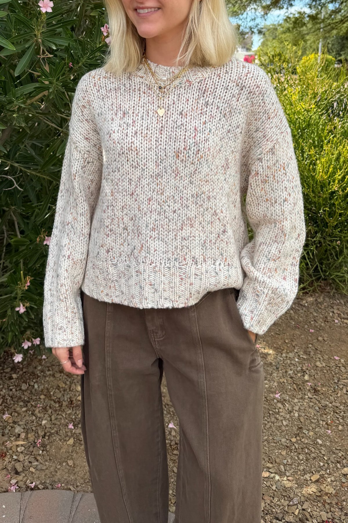 A woman wearing a cream-colored, hand-knit long-sleeve sweater with subtle multicolor flecks, paired with brown pants, standing outdoors in front of green foliage.