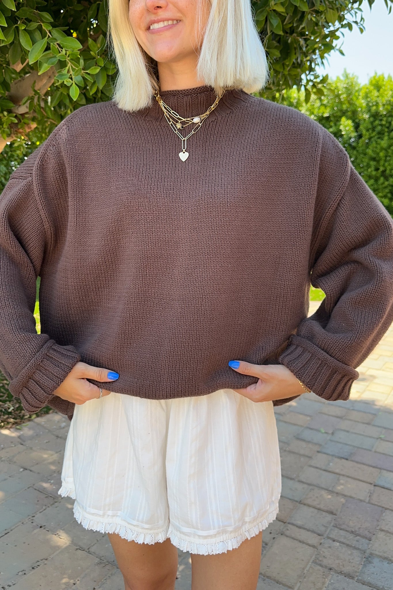 A woman wearing a brown ribbed knit mock neck sweater and white shorts, standing outdoors on a paved walkway with green foliage in the background.