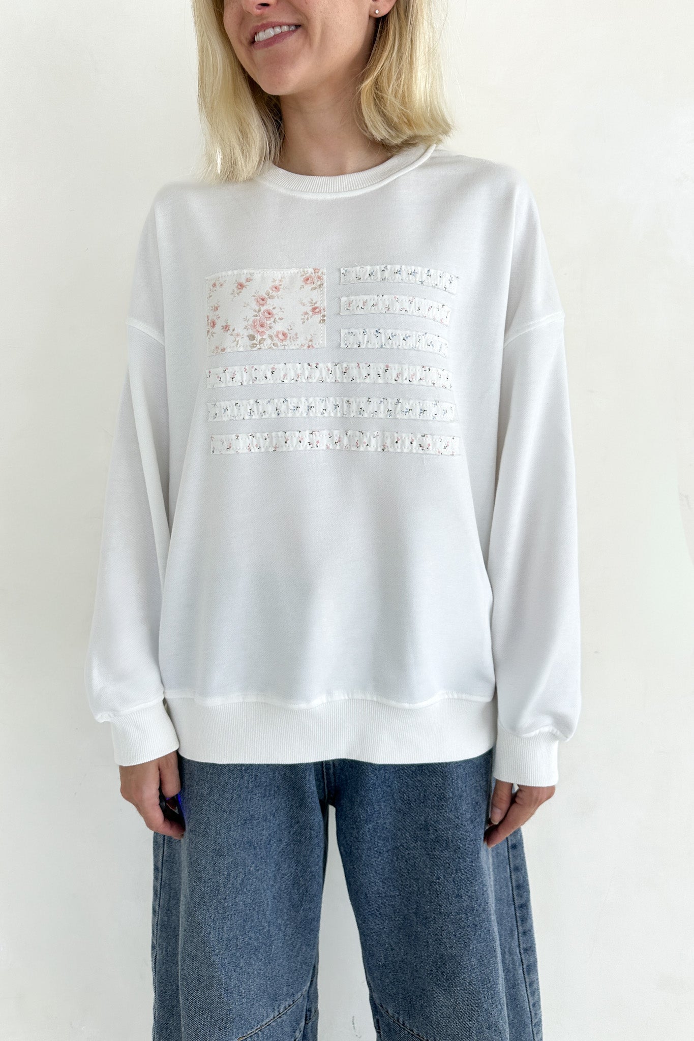 Person wearing a white sweatshirt with lace details and blue jeans on a plain background