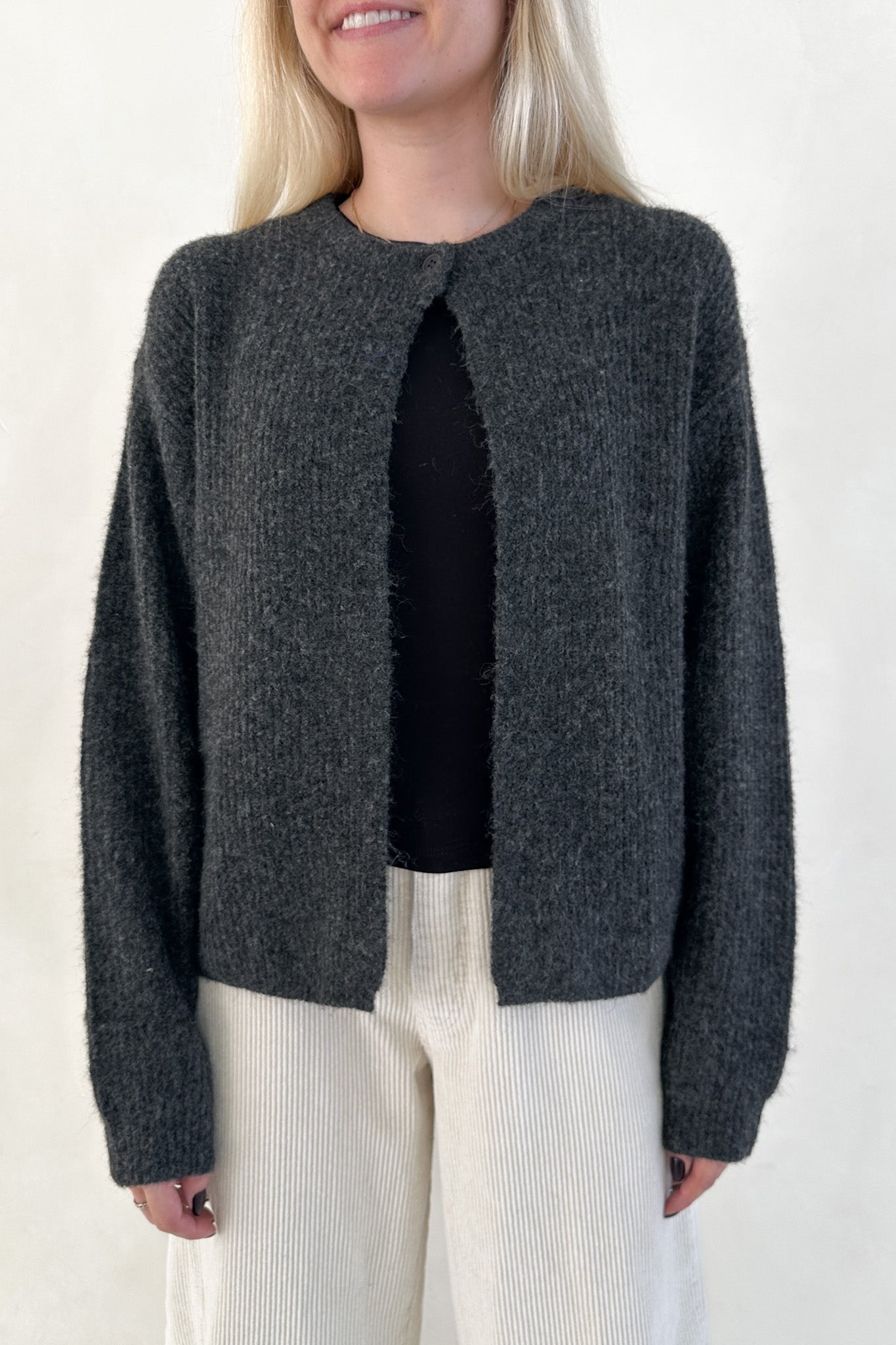 Person wearing a dark gray cardigan over a black top and white pants on a plain background