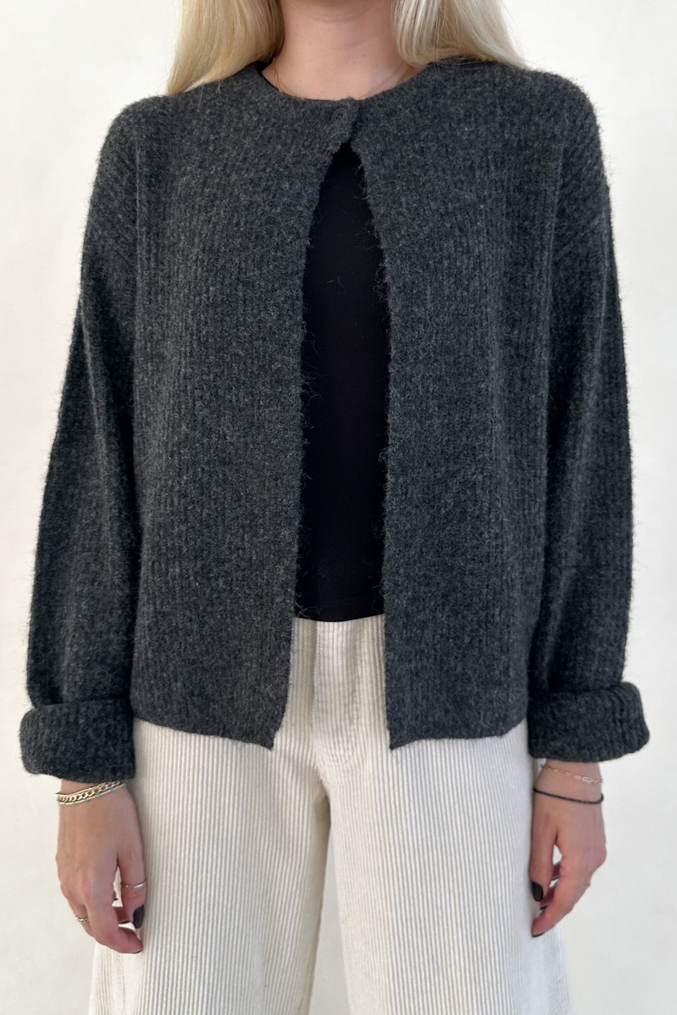 Person wearing a dark gray textured jacket over a black top with white pants on a plain background