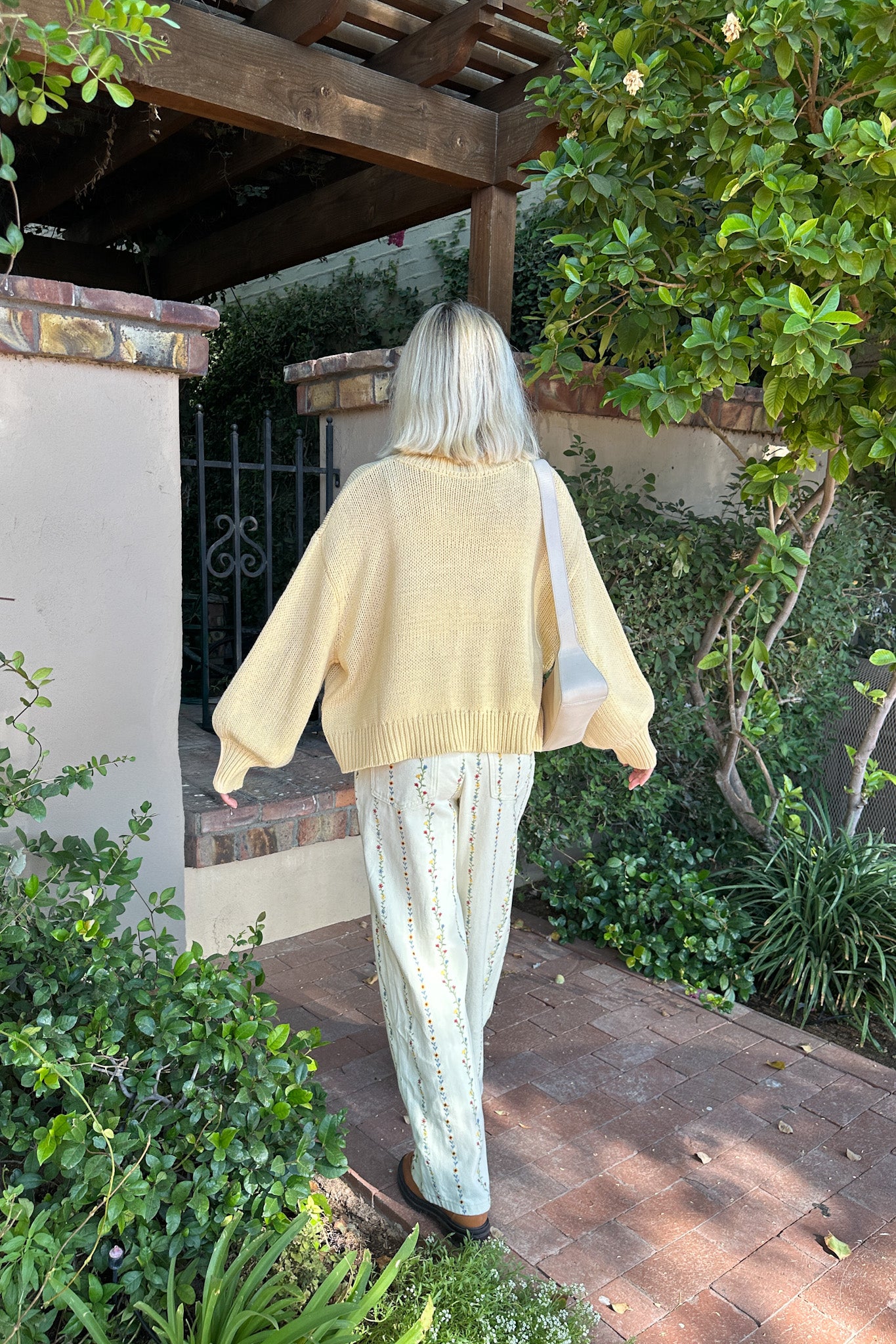 Person wearing a beige cardigan and light-colored pants walking outdoors near greenery.