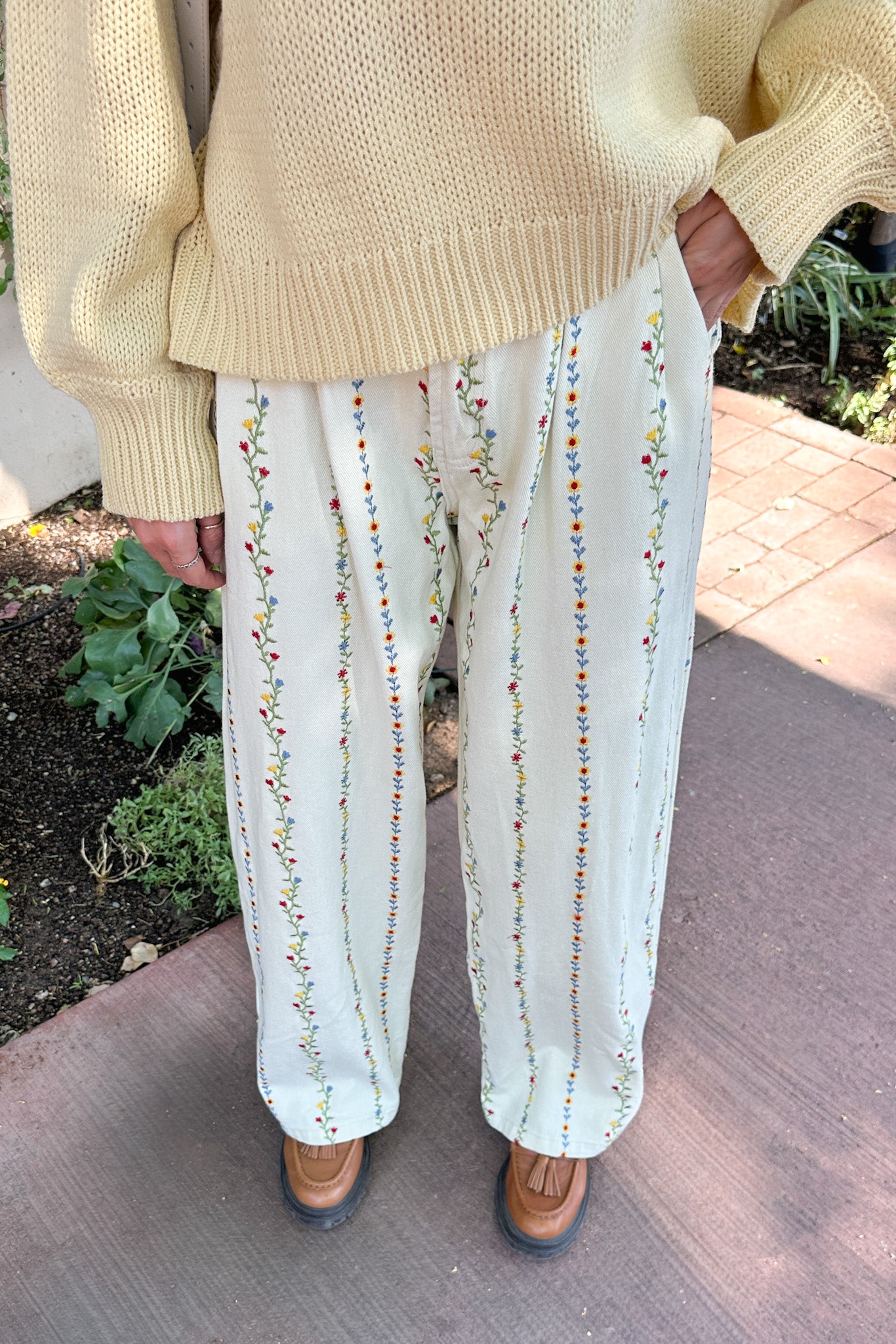 Person wearing a beige sweater and white pants with colorful embroidery, standing on a sidewalk.