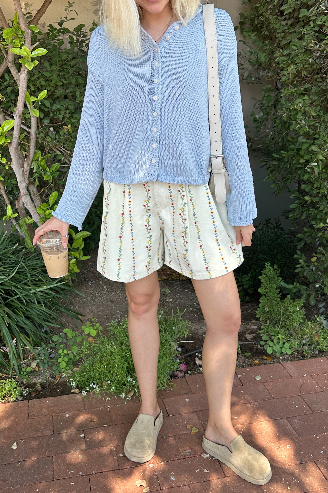 Person wearing a light blue shirt and patterned shorts, holding a drink, standing outdoors.