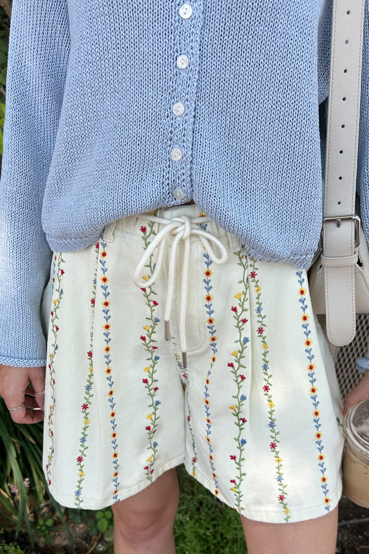 Person wearing a blue cardigan and white shorts with colorful embroidery, standing outdoors.