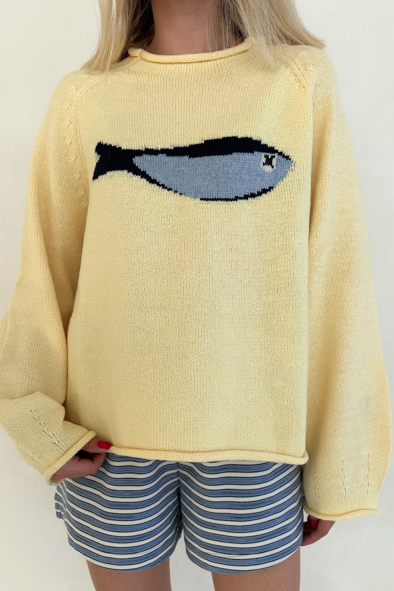 Person wearing a yellow sweater with a fish design and striped shorts on a plain background