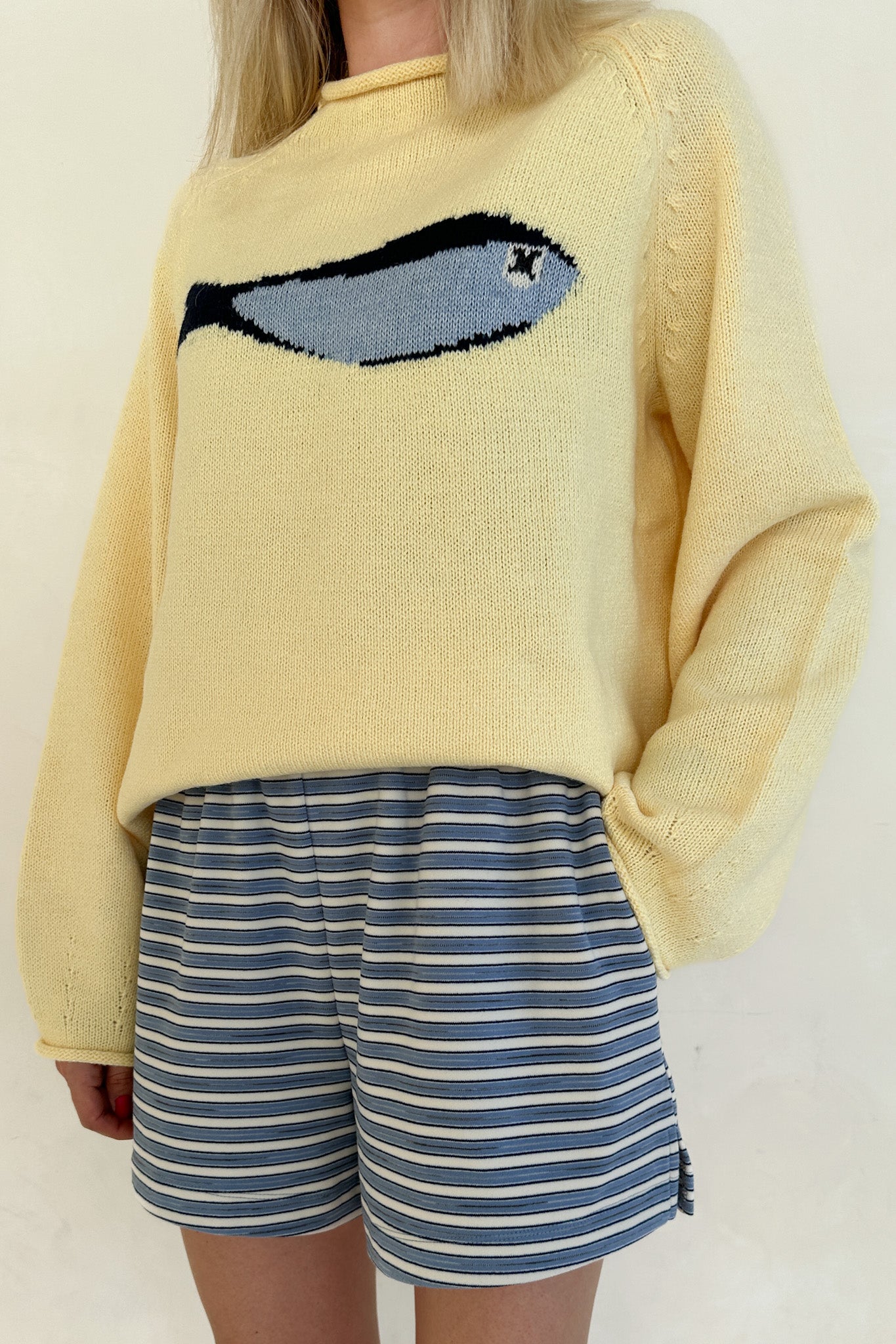 Person wearing a yellow sweater with a fish design and blue striped shorts on a plain background