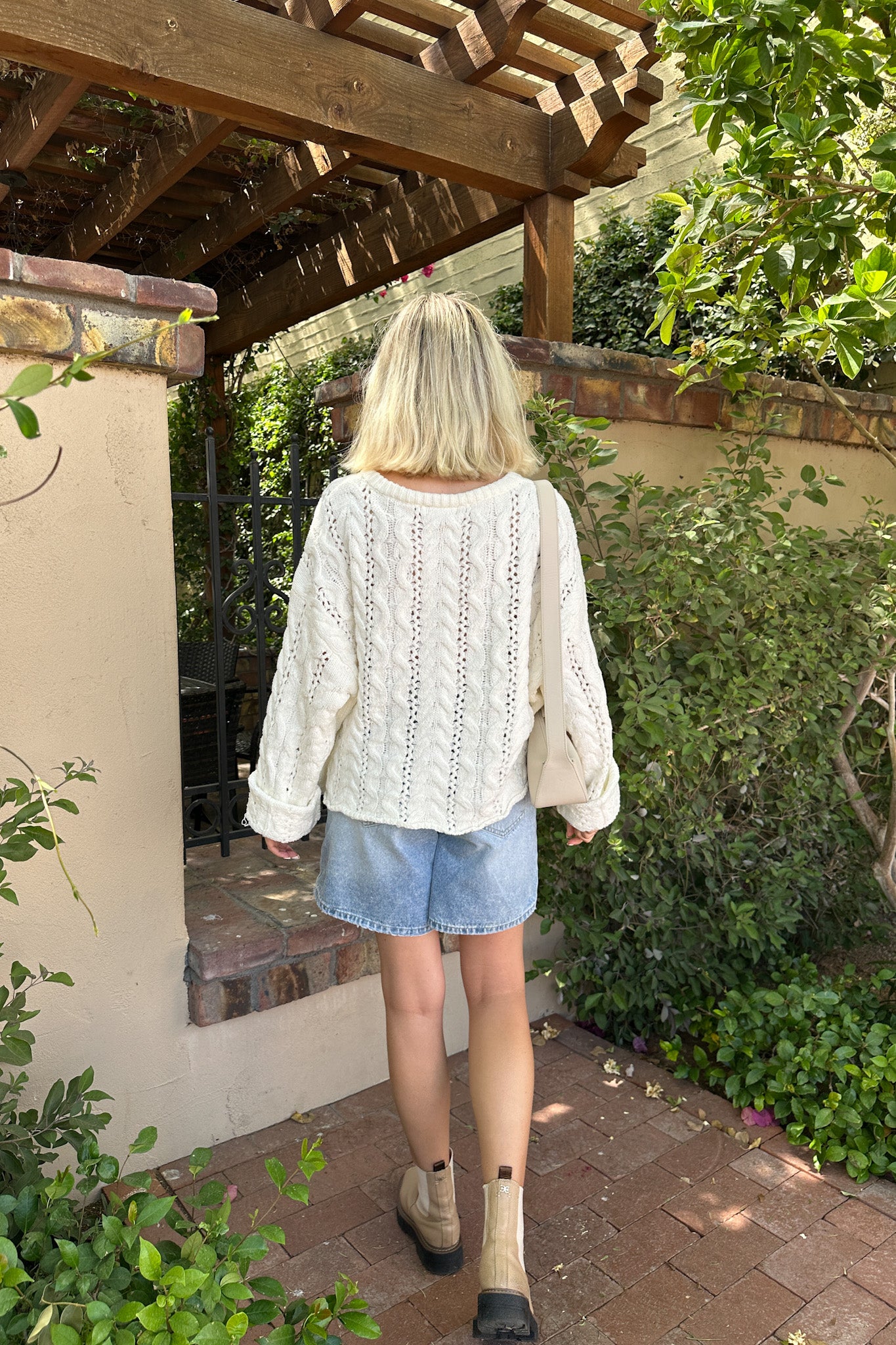 Person wearing a white lace top and denim shorts standing under a wooden pergola with greenery around.