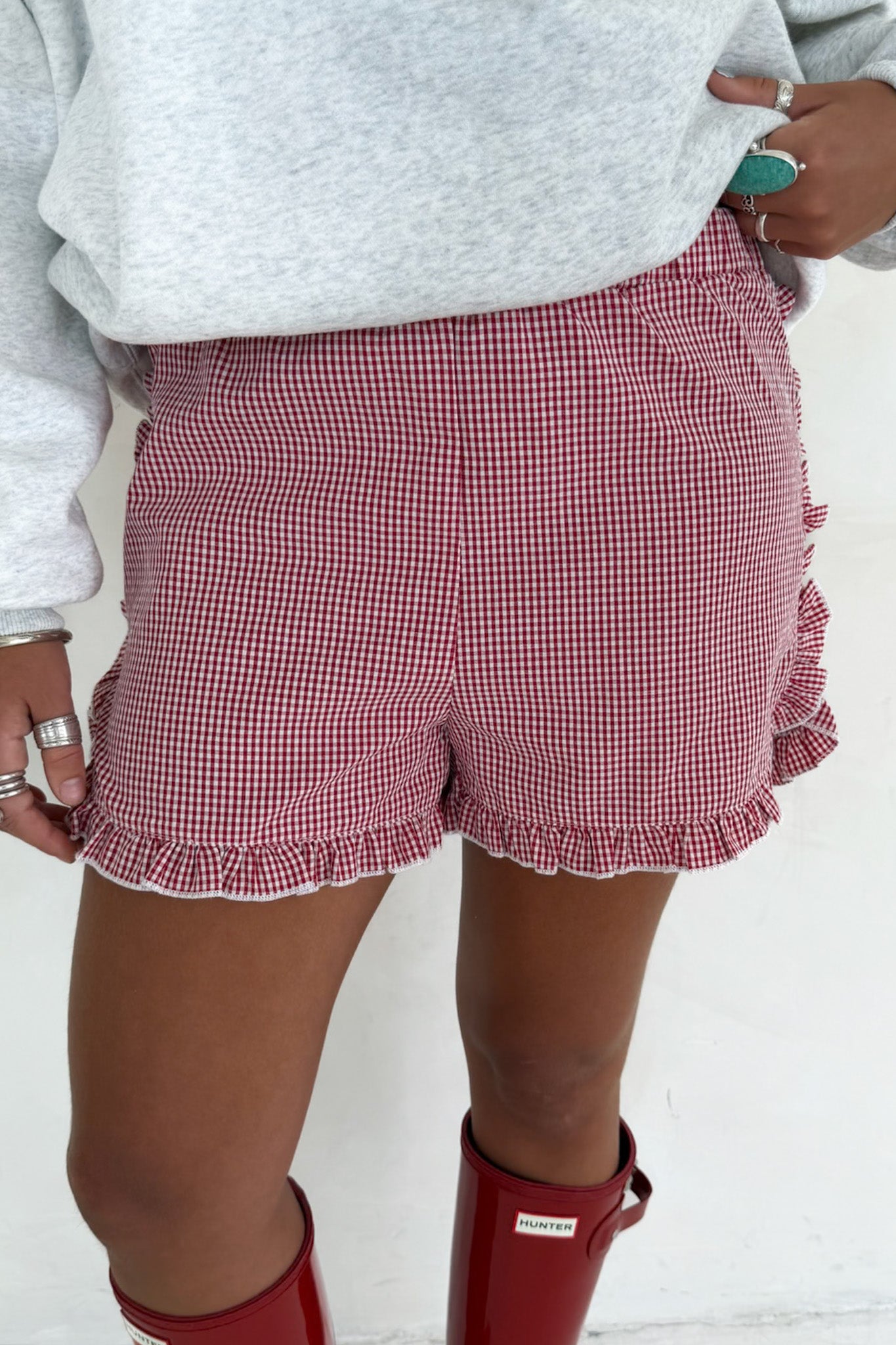 A person wearing red and white gingham check ruffle shorts, a light gray sweatshirt, and red rain boots.