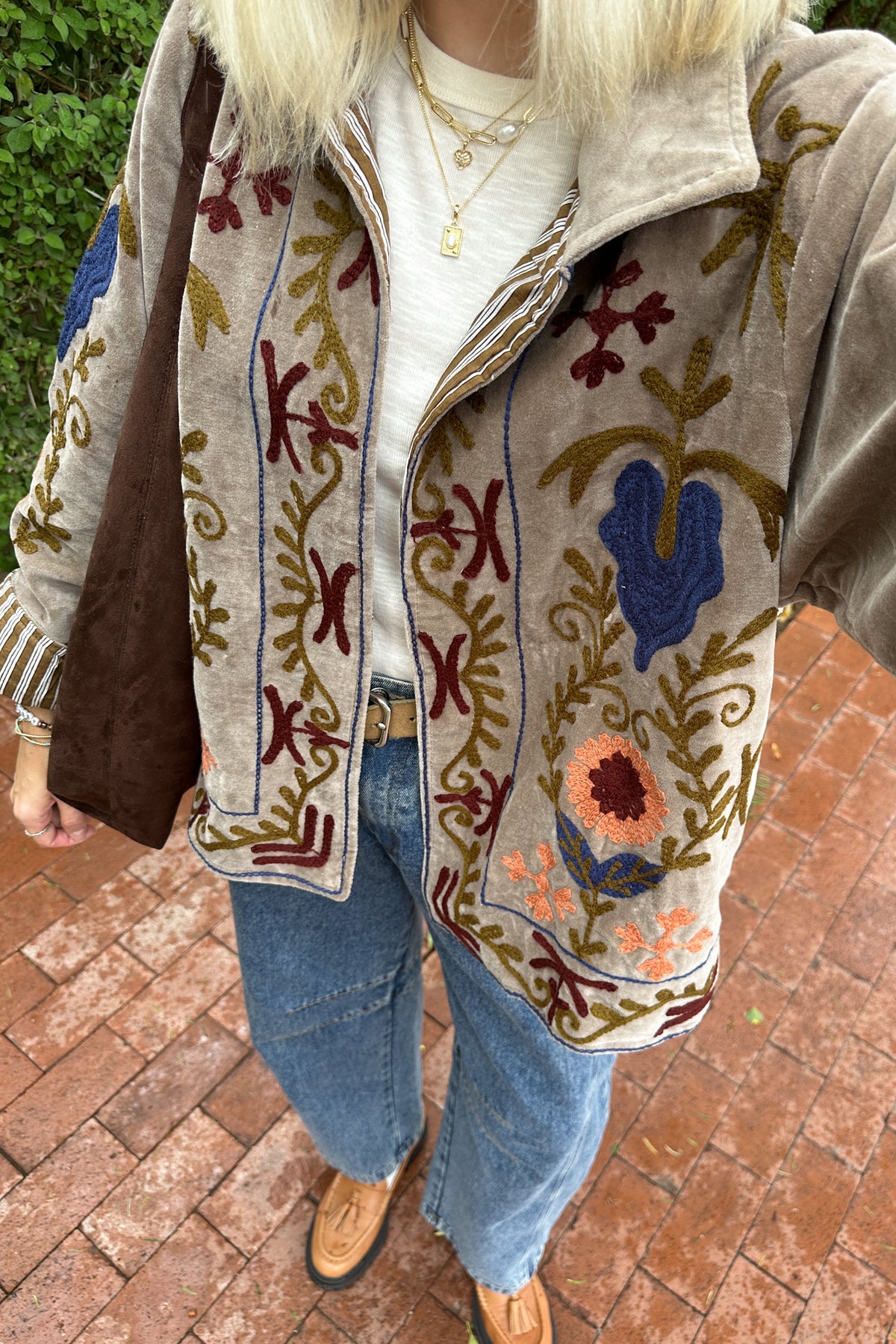A person wearing a beige embroidered jacket with floral and geometric patterns in blue, brown, and orange, paired with a white top, blue jeans, and brown shoes, standing on a brick patio.
