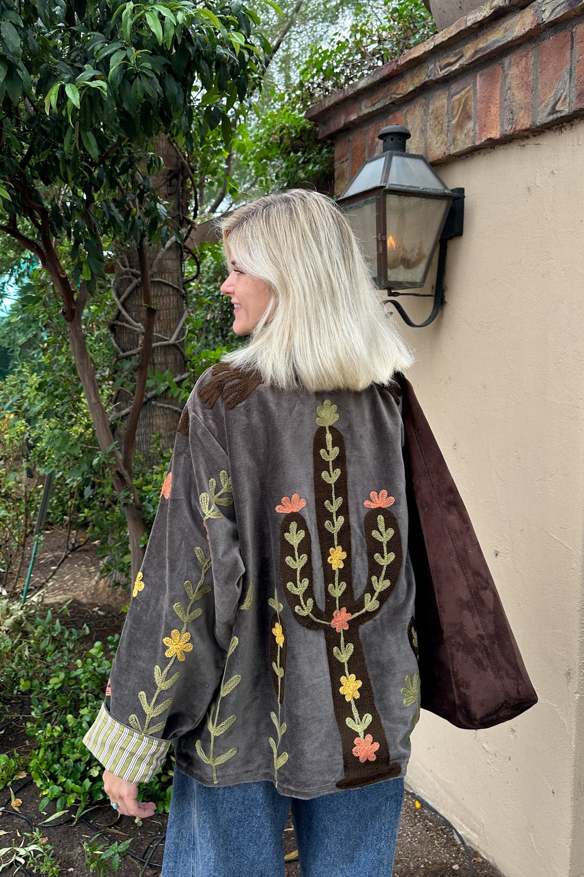 A woman with blonde hair stands outdoors in a garden setting, wearing a dark gray suede jacket with a large embroidered cactus and flowers on the back, paired with blue jeans.