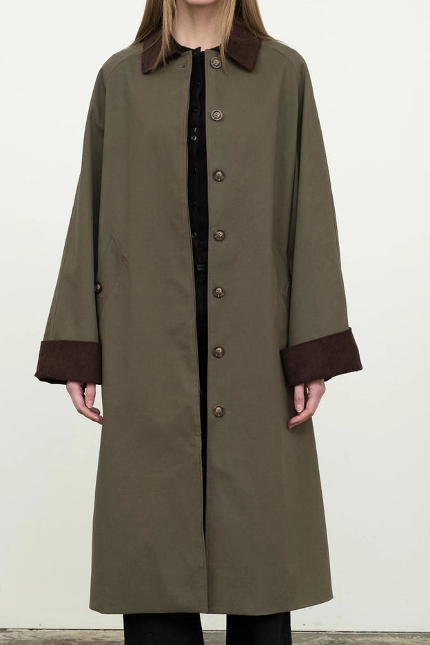 Person wearing a long olive green coat with brown trim against a white background