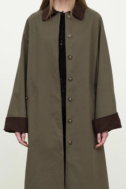Person wearing a long olive green coat with brown trim against a white background