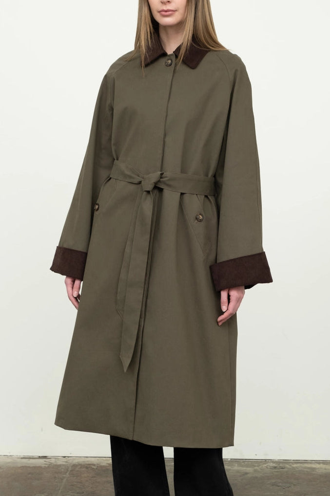 Woman wearing a long olive green coat with brown trim against a white background