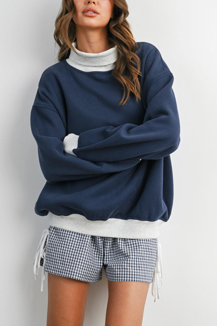 A woman modeling an oversized navy blue sweatshirt with a white mock neck insert and white drawstring ties, paired with blue and white gingham shorts, against a plain light gray background.