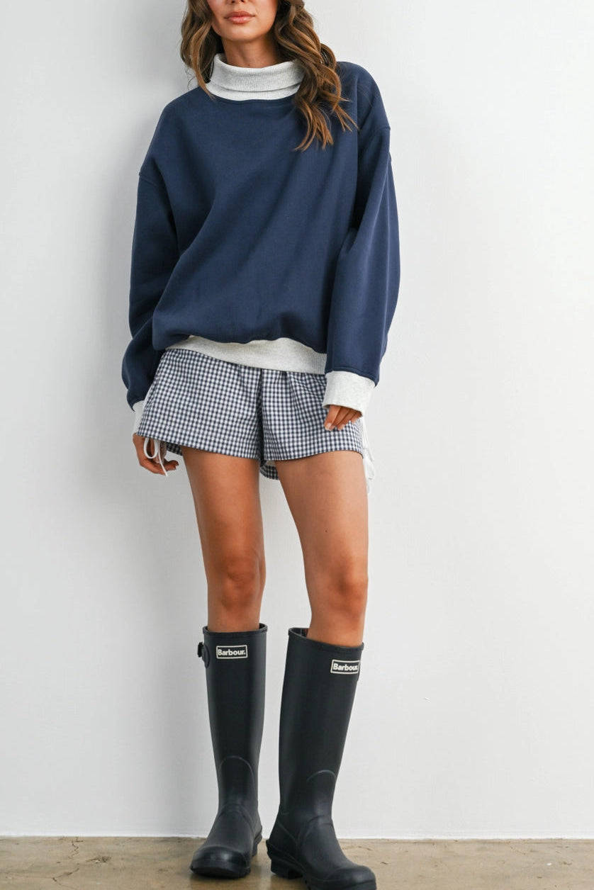 A woman stands against a plain white wall wearing a navy blue sweatshirt over a white turtleneck, blue-and-white gingham shorts, and black rain boots with visible Hunter branding.