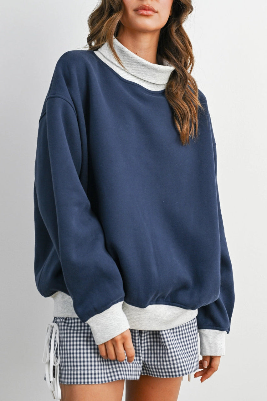 A woman wearing an oversized navy blue sweatshirt with a contrasting white mock neck, ribbed hem, and cuffs, paired with blue and white gingham shorts, against a light gray studio background.