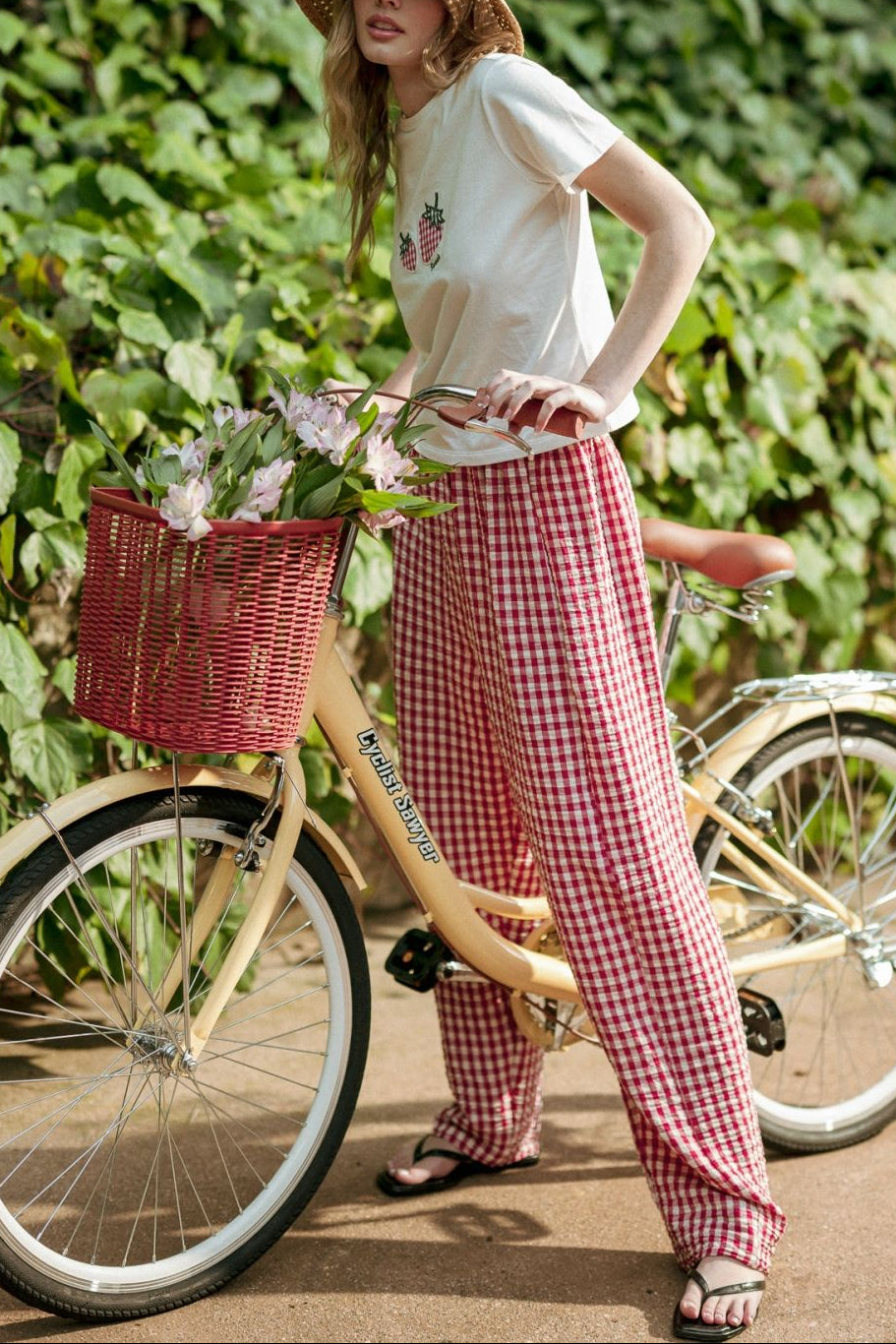 Woman in a white shirt and red checkered pants standing next to a bicycle with a basket, against a green leafy background.